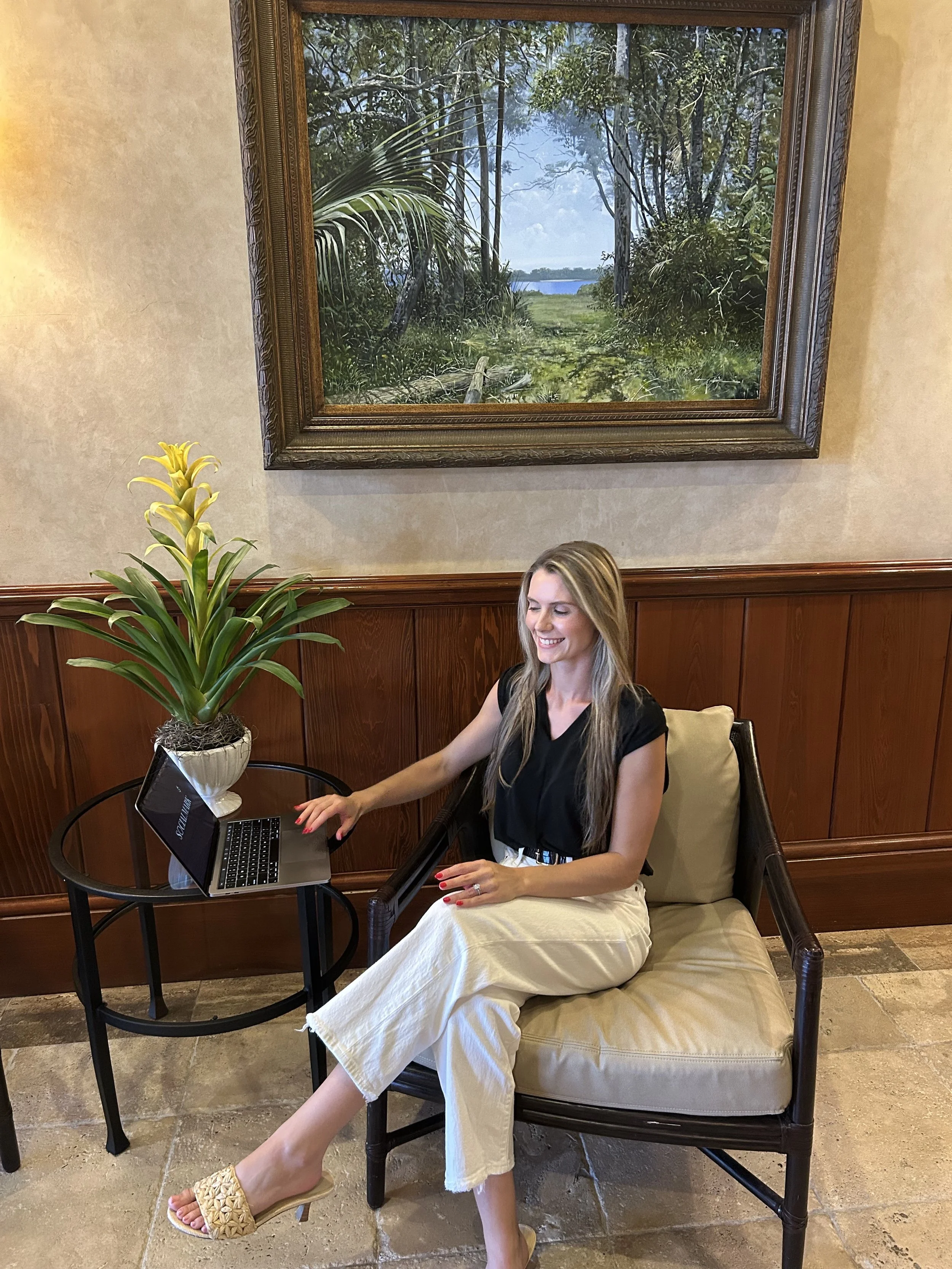 A woman with long blond hair sitting in a cushioned chair, smiling while using a laptop on a round black table. She is wearing an all-black top, white wide-leg pants, and woven beige sandals. To her left, there is a tall potted plant with yellow flowers. Behind her, a large framed painting of a green wooded landscape with a path leading to water is hung on a beige wall with wood paneling. The floor is made of stone tiles.