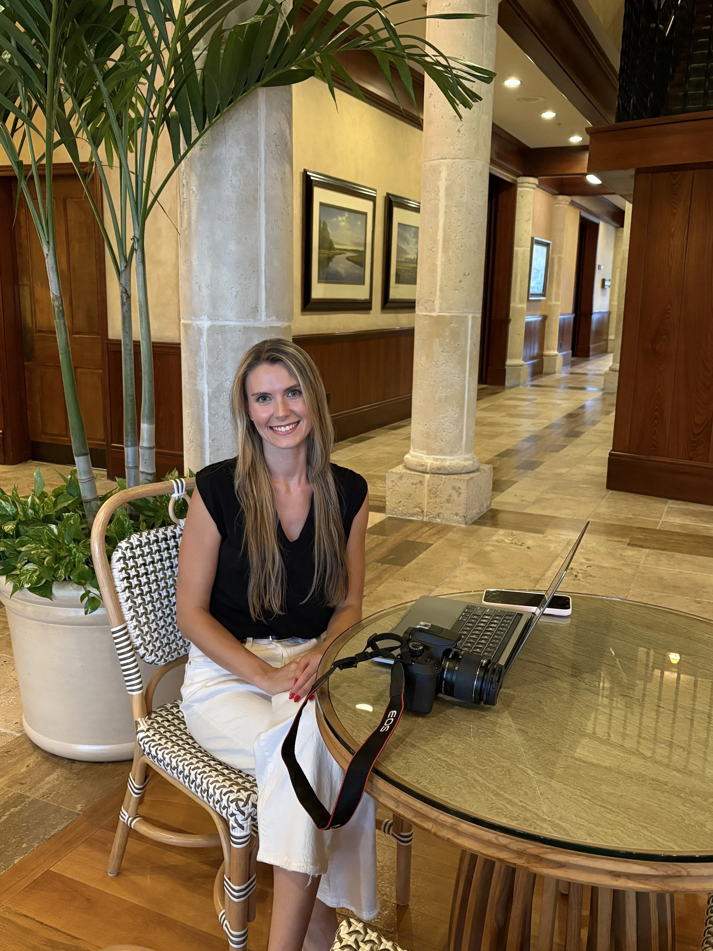 A woman sitting at a round wooden table in a hotel lobby or restaurant, smiling, with a DSLR camera, laptop, and smartphone on the table. She is dressed in a black top and white pants, with long blonde hair. There is a large potted plant nearby, and the background features stone columns, framed landscape paintings, and warm wooden paneling.