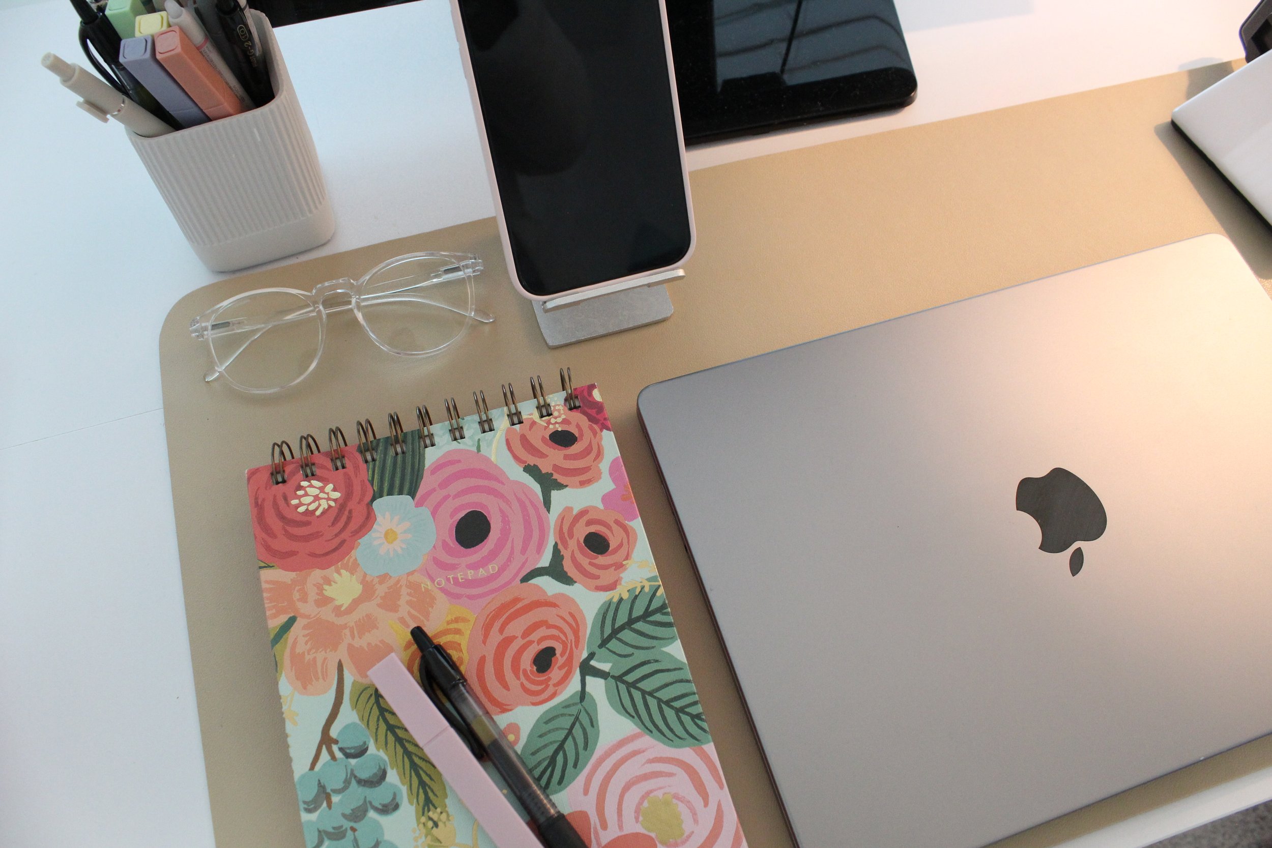 A desk with a white container holding pens and markers, a pair of clear eyeglasses, a floral patterned notebook, a smartphone on a stand, a silver laptop, a closed laptop, and a white charging cable.