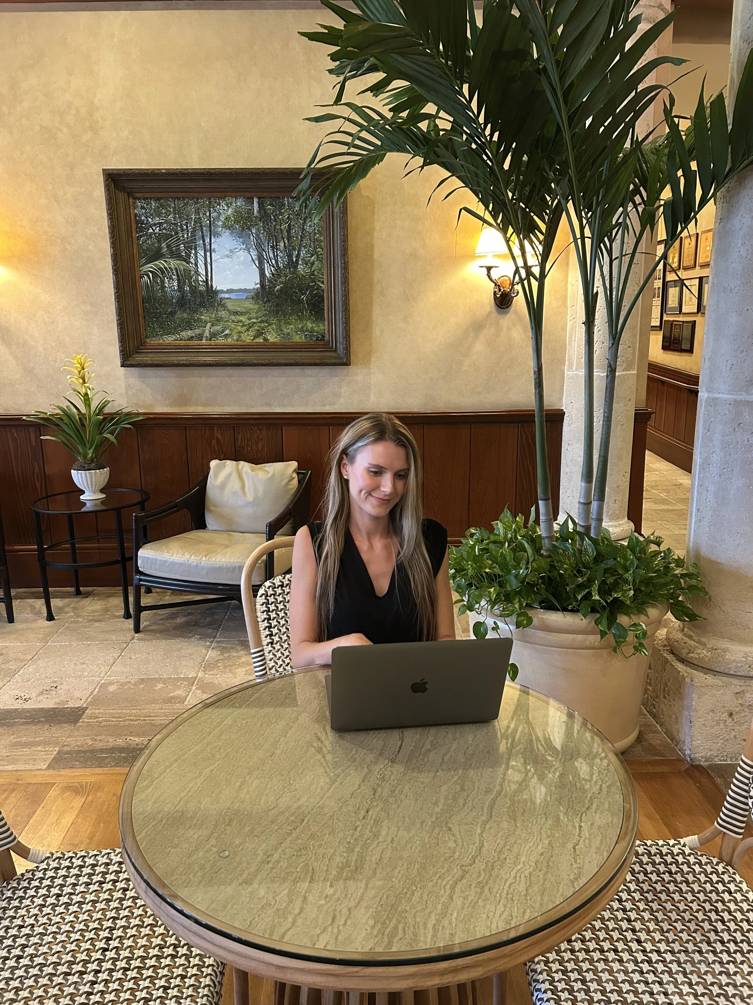 Woman sitting at a glass-top table with a laptop in a restaurant or hotel lobby, decorated with large potted plants and framed artwork on the wall.