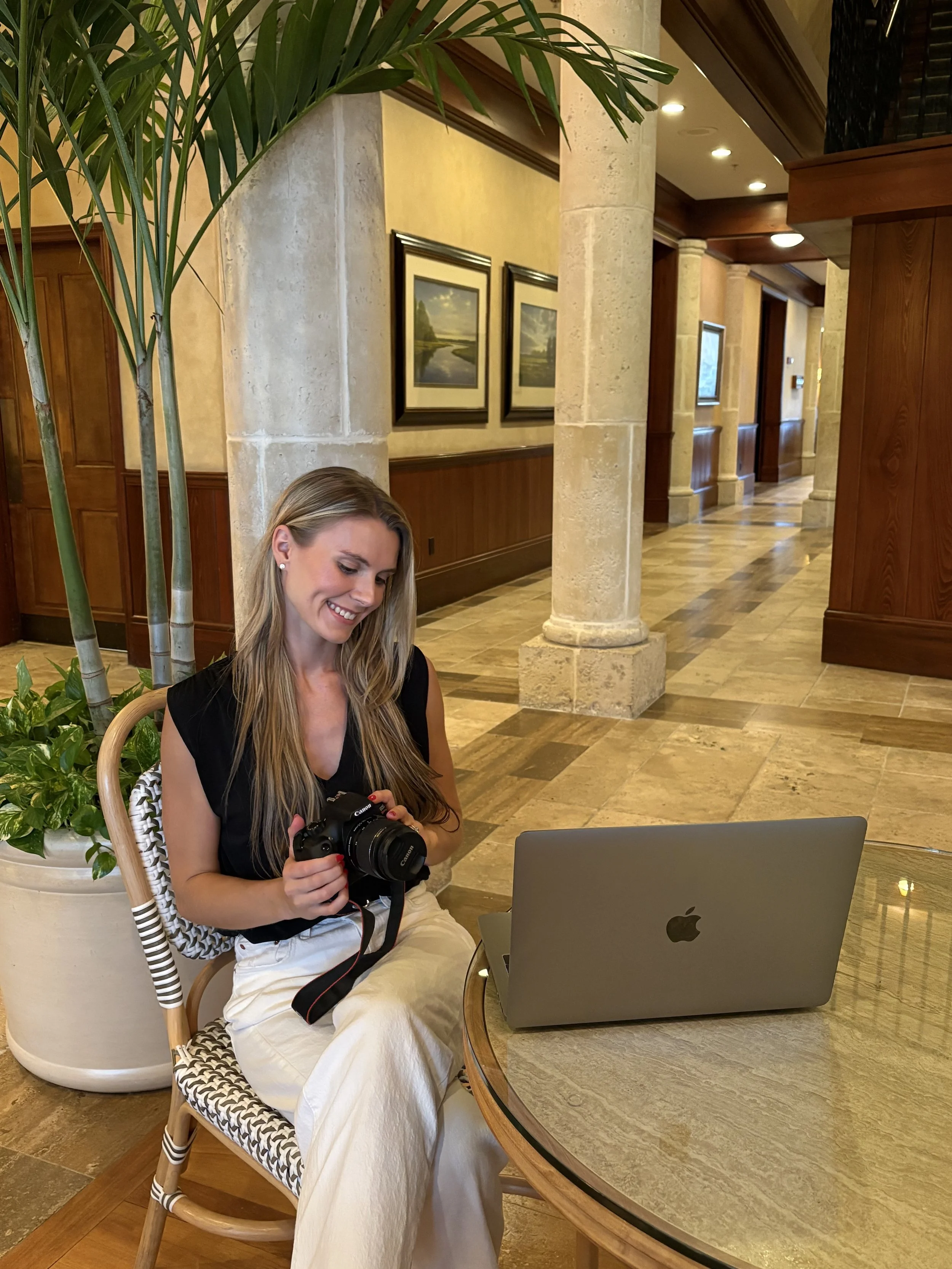 A woman with long blonde hair, wearing a black top and beige pants, is sitting at a round wooden table in a warmly lit hotel lobby or lounge. She is smiling and looking at a DSLR camera in her hands. In front of her on the table is an open silver MacBook laptop. Behind her is a large potted plant, stone columns, and framed landscape paintings on the wall.