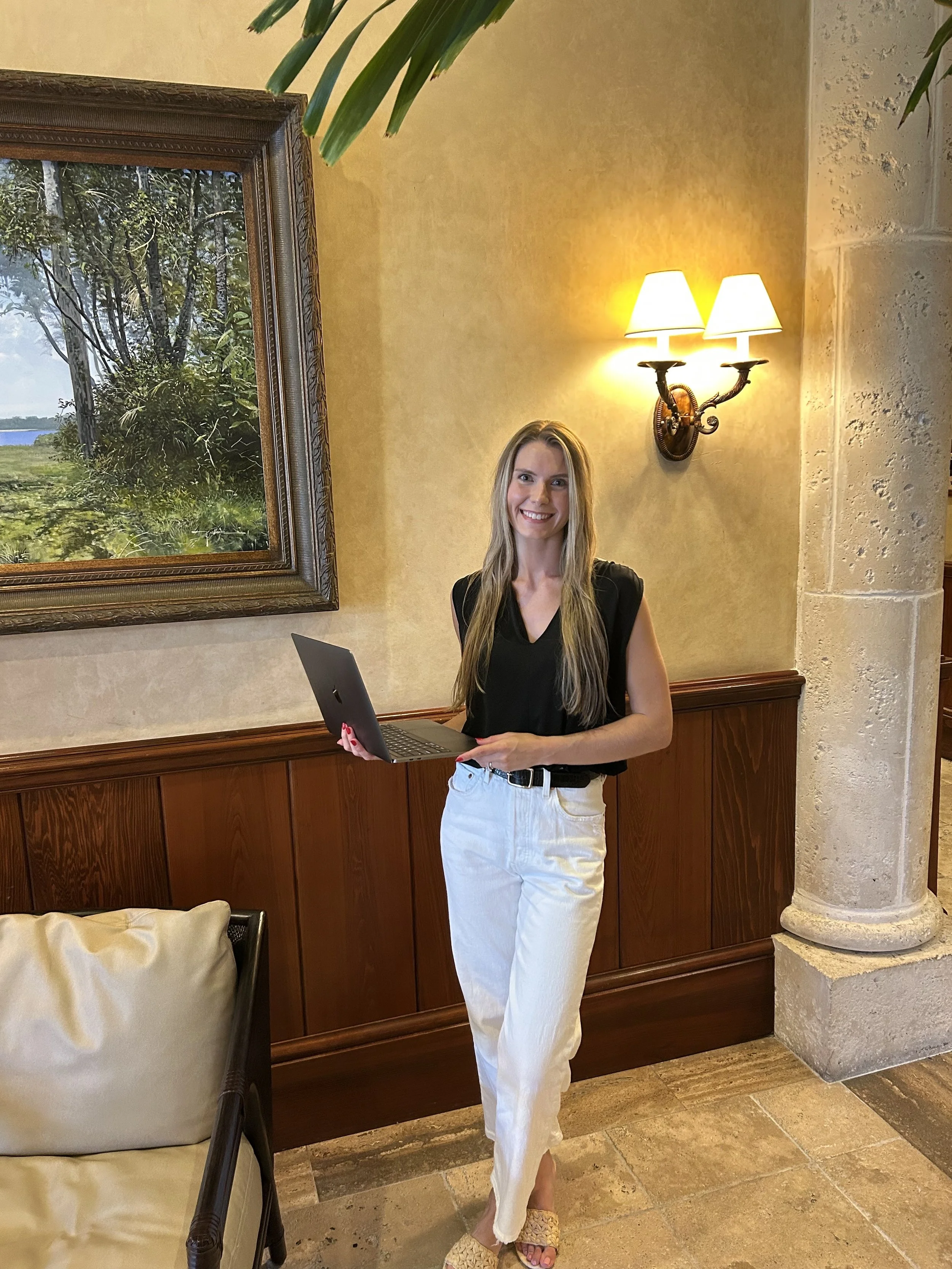 A woman with long blonde hair, wearing a black sleeveless top, white pants, and beige woven sandals, standing indoors next to a beige pillow on a dark wicker chair, holding a laptop. Behind her, there is a framed nature painting, a wall sconce with two lamps, and textured beige walls with stone accents.