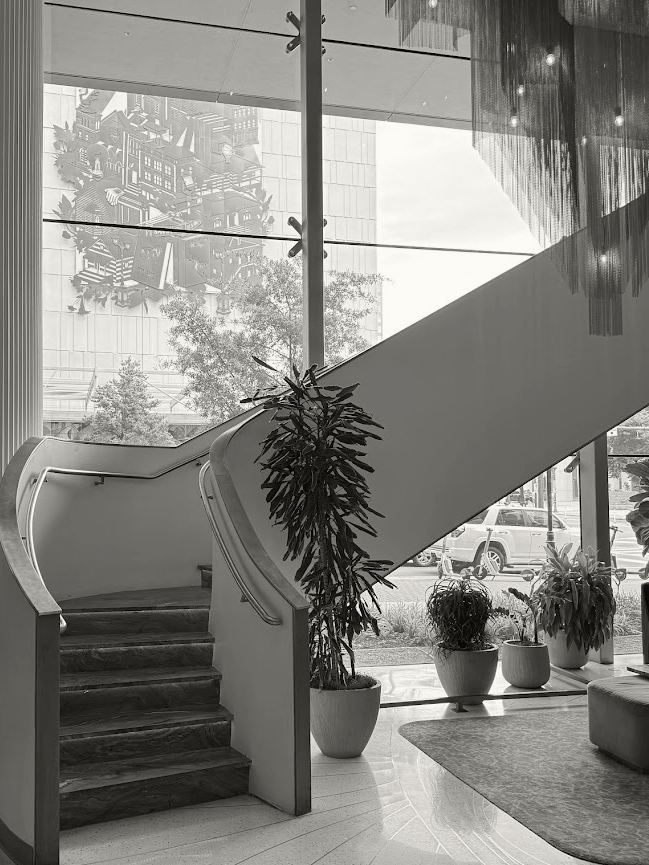 Black and white image of a staircase, potted plant and chandelier. Through the window is a mural painted on the side of a wall.