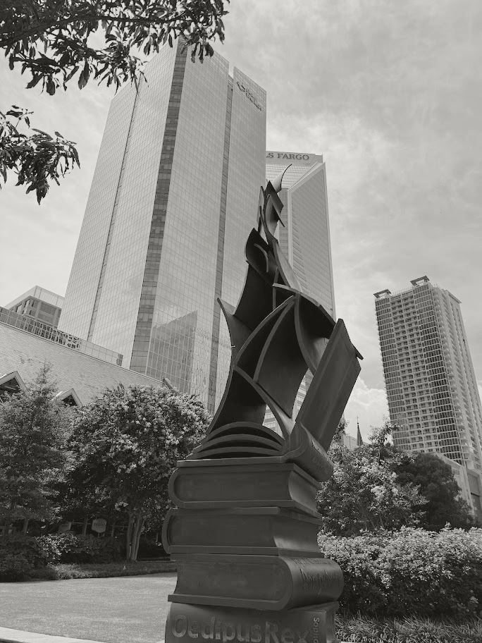 A sculpture of a sailing ship made of metal, mounted on top of large stacked books, located in an outdoor urban park with tall modern buildings in the background.