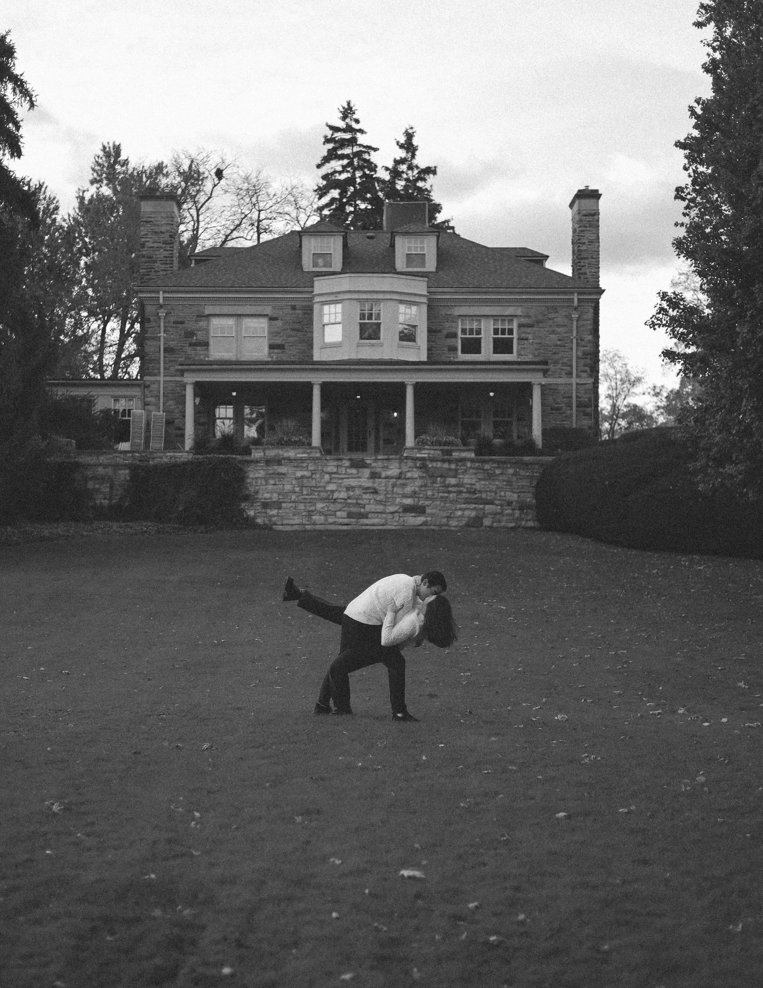 A black and white photograph of a man and woman dancing on a lawn in front of a large, historic house with a stone facade and multiple chimneys. The man is wearing a white shirt and dark pants, while the woman is wearing dark clothing. They are close