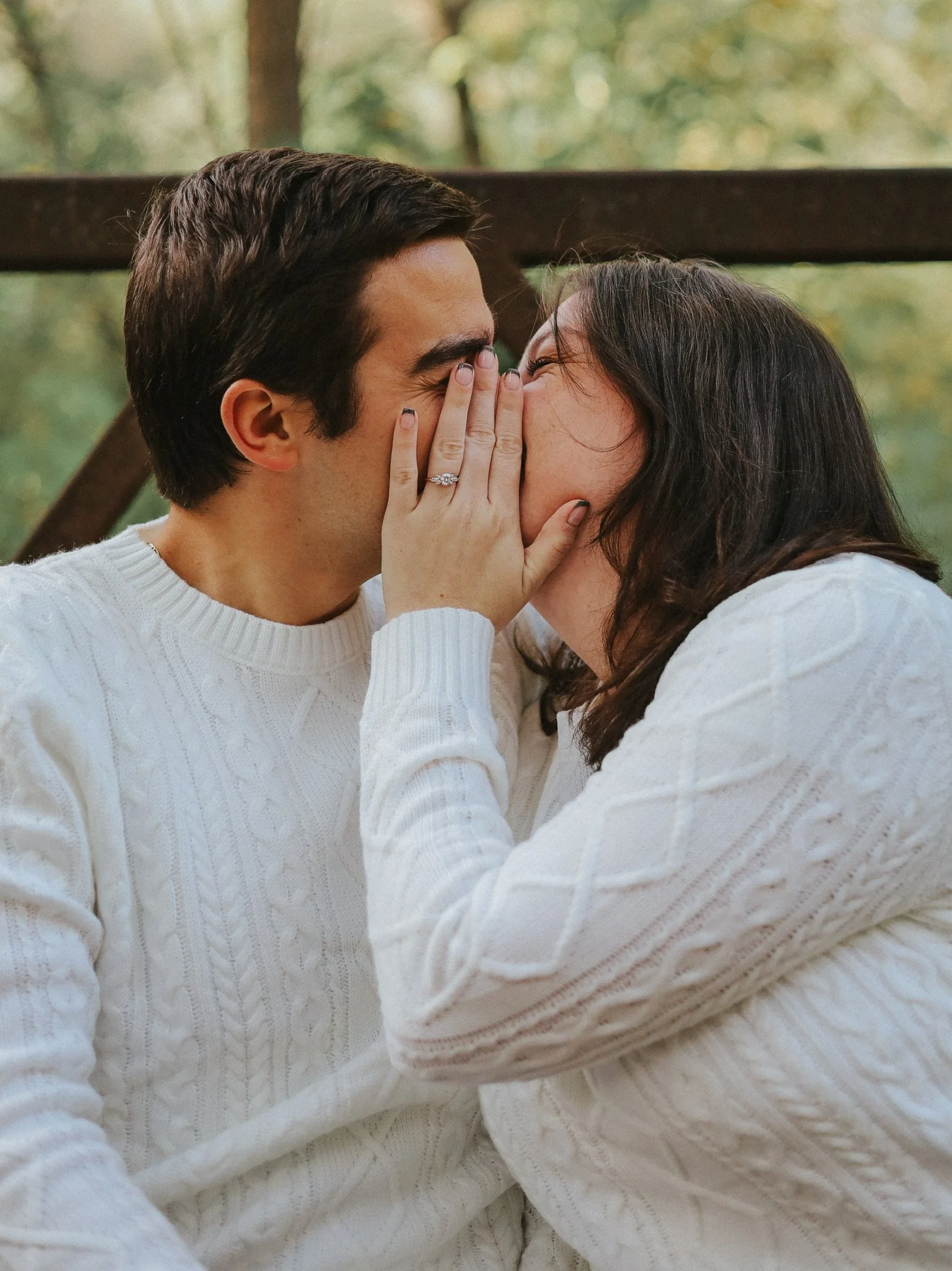 A couple sharing a kiss, wearing white sweaters, with a blurred outdoor background.