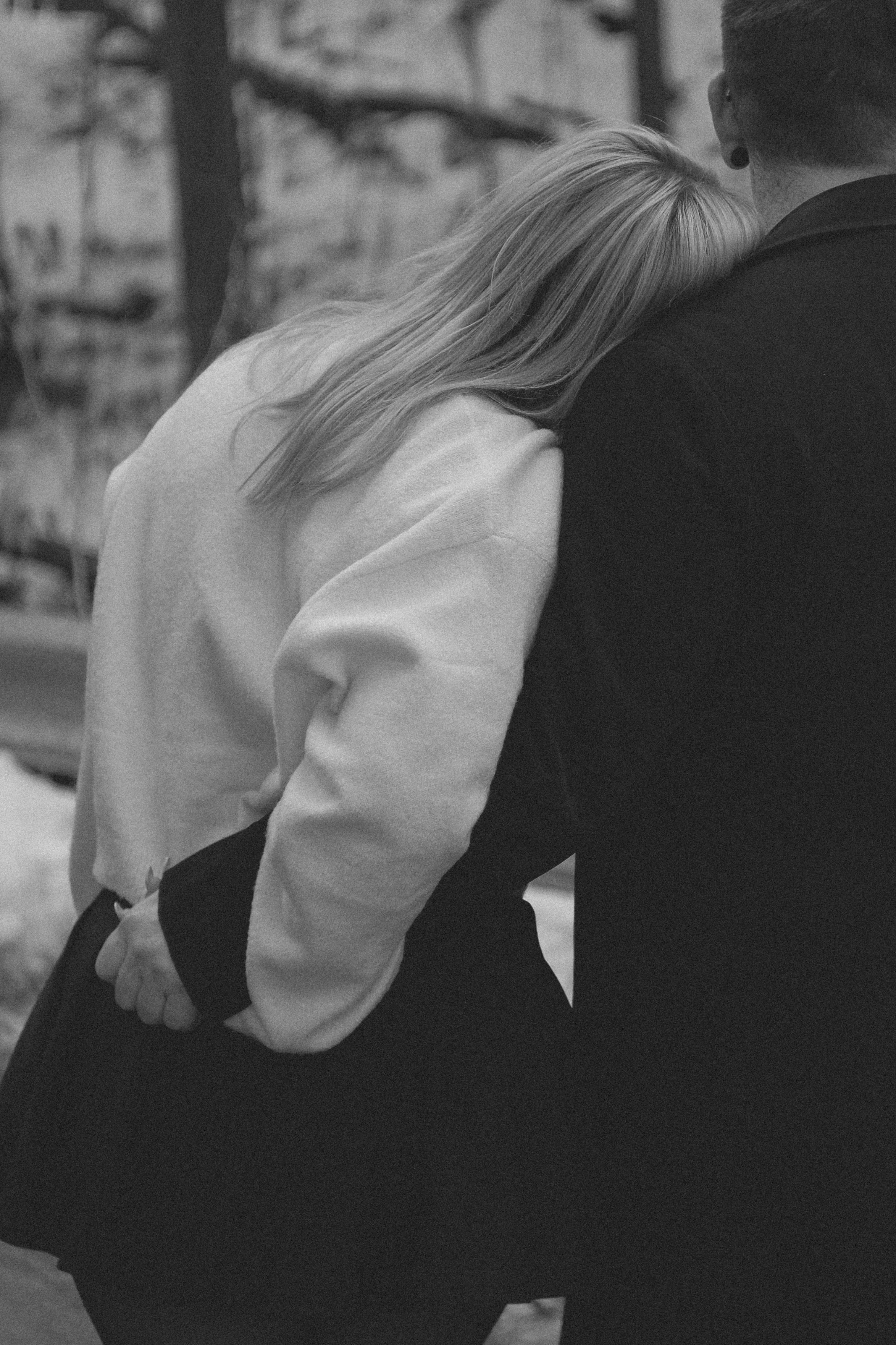 A black and white photo showing a woman resting her head on a man's shoulder, both are wearing dark clothing and standing outdoors with trees in the background.