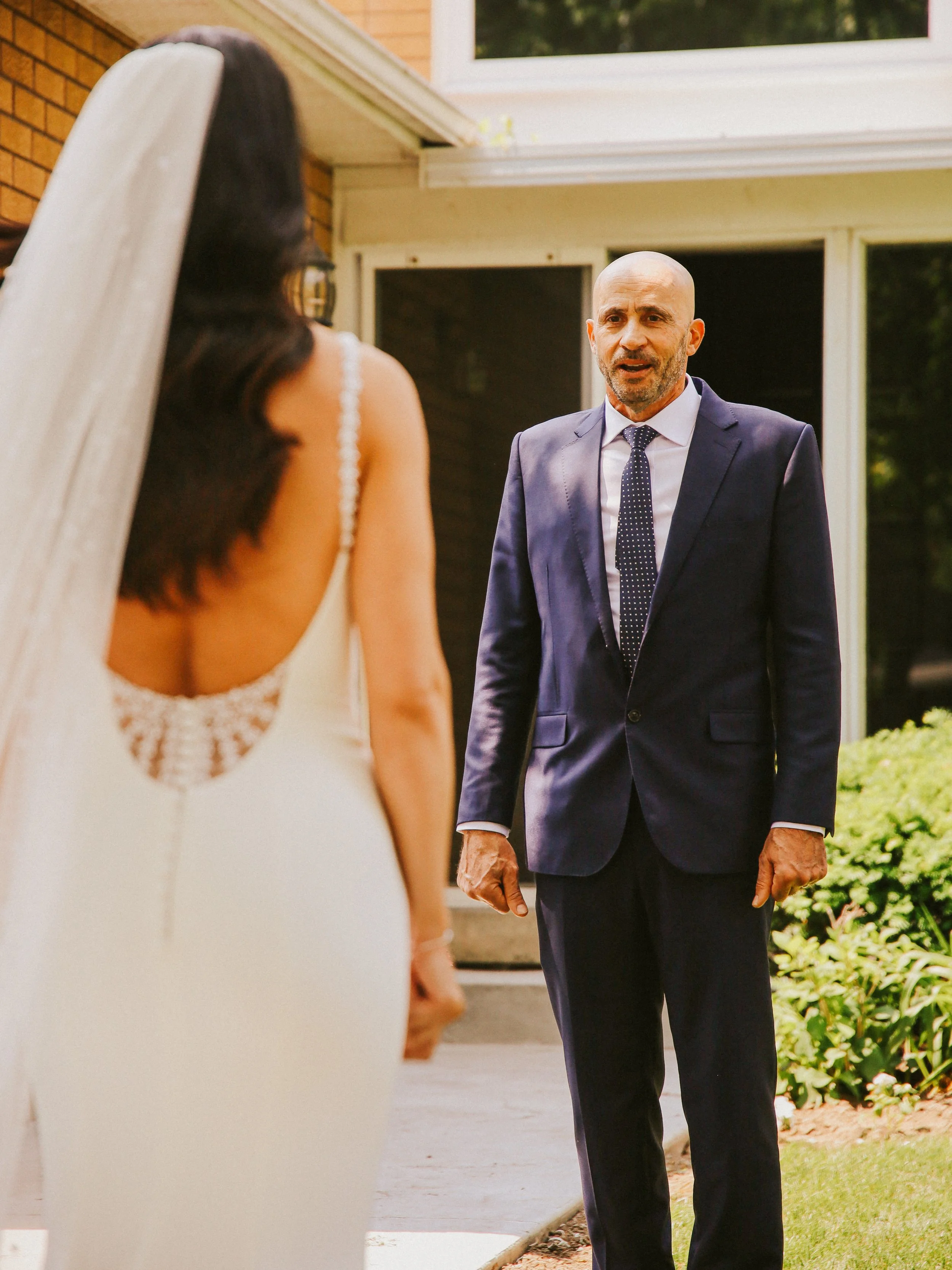 An older man with a bald head, wearing a suit and a polka dot tie, is standing outside in front of a house, looking at a woman in a wedding dress with a veil.