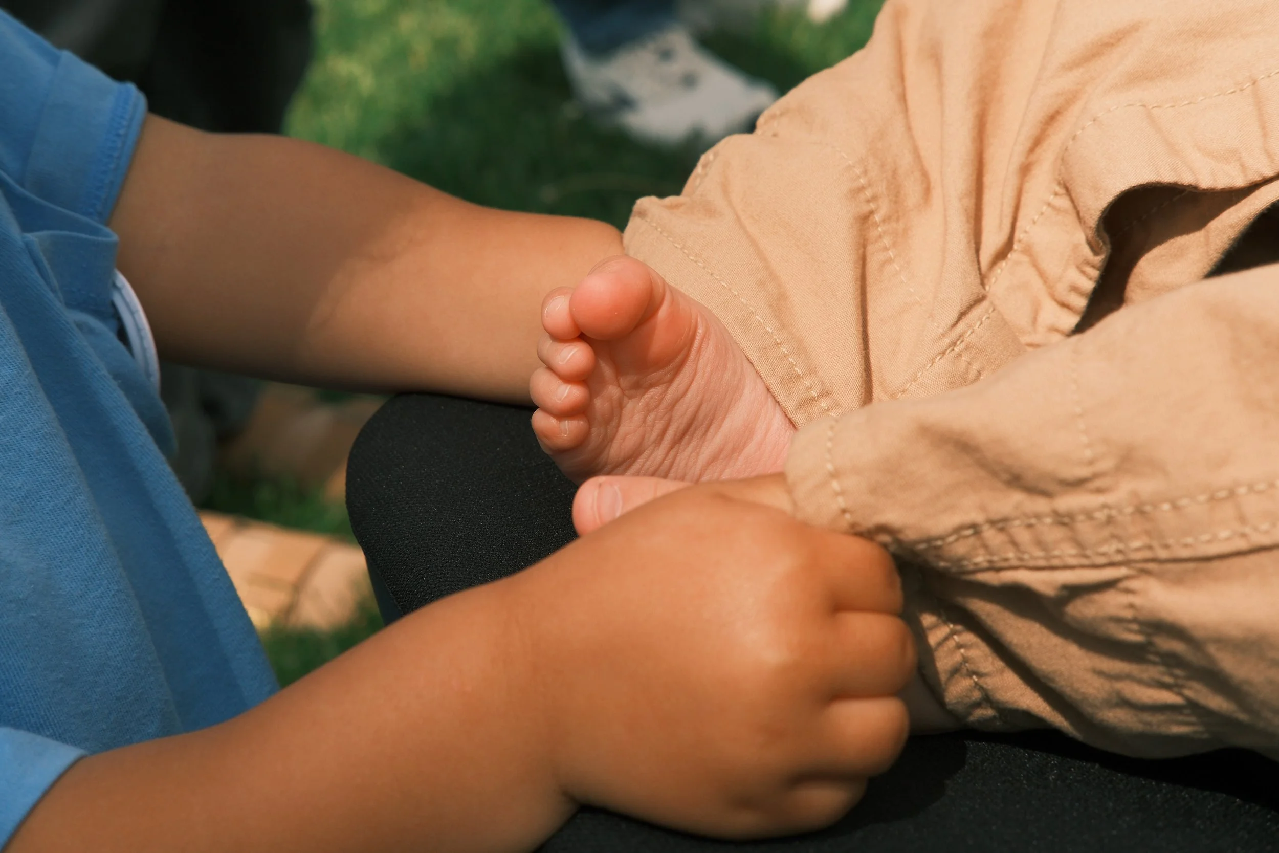Close-up of a child's arm and hand, with the child making a fist, resting against an adult's arm, who is wearing a tan shirt. The background is blurred, showing green grass.
