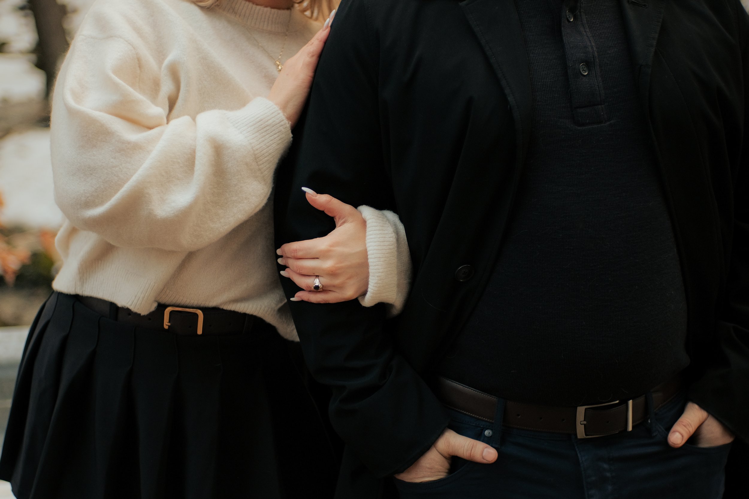 A close-up of a woman with a beige sweater holding hands on a man's arm, who is wearing a black jacket and black shirt, with a stern gesture of touching his belt.