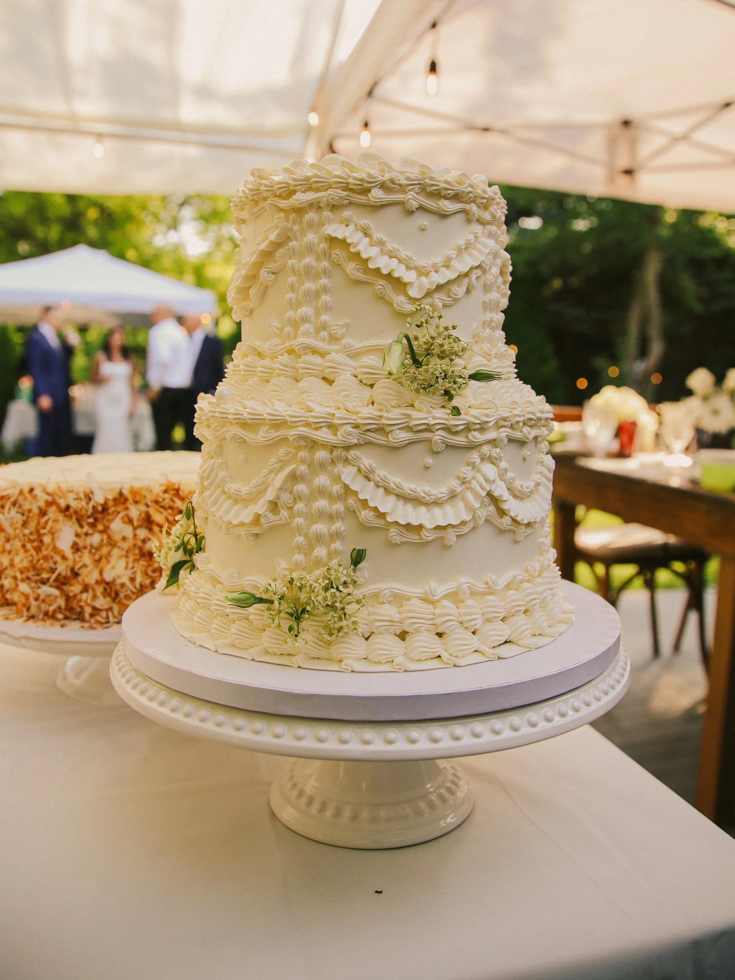 A tall, white wedding cake decorated with intricate piping, small white flowers, and greenery, placed on a stand at an outdoor wedding reception.