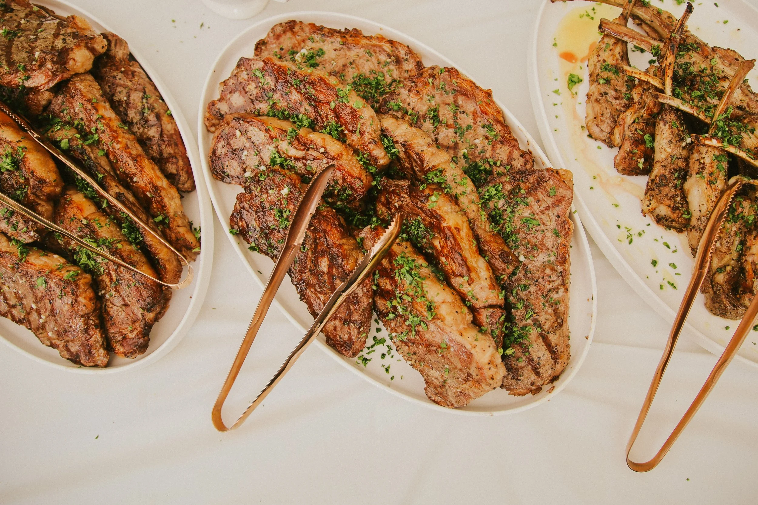 Close-up of three white plates of grilled meats garnished with chopped herbs, with tongs resting on top of the meats.