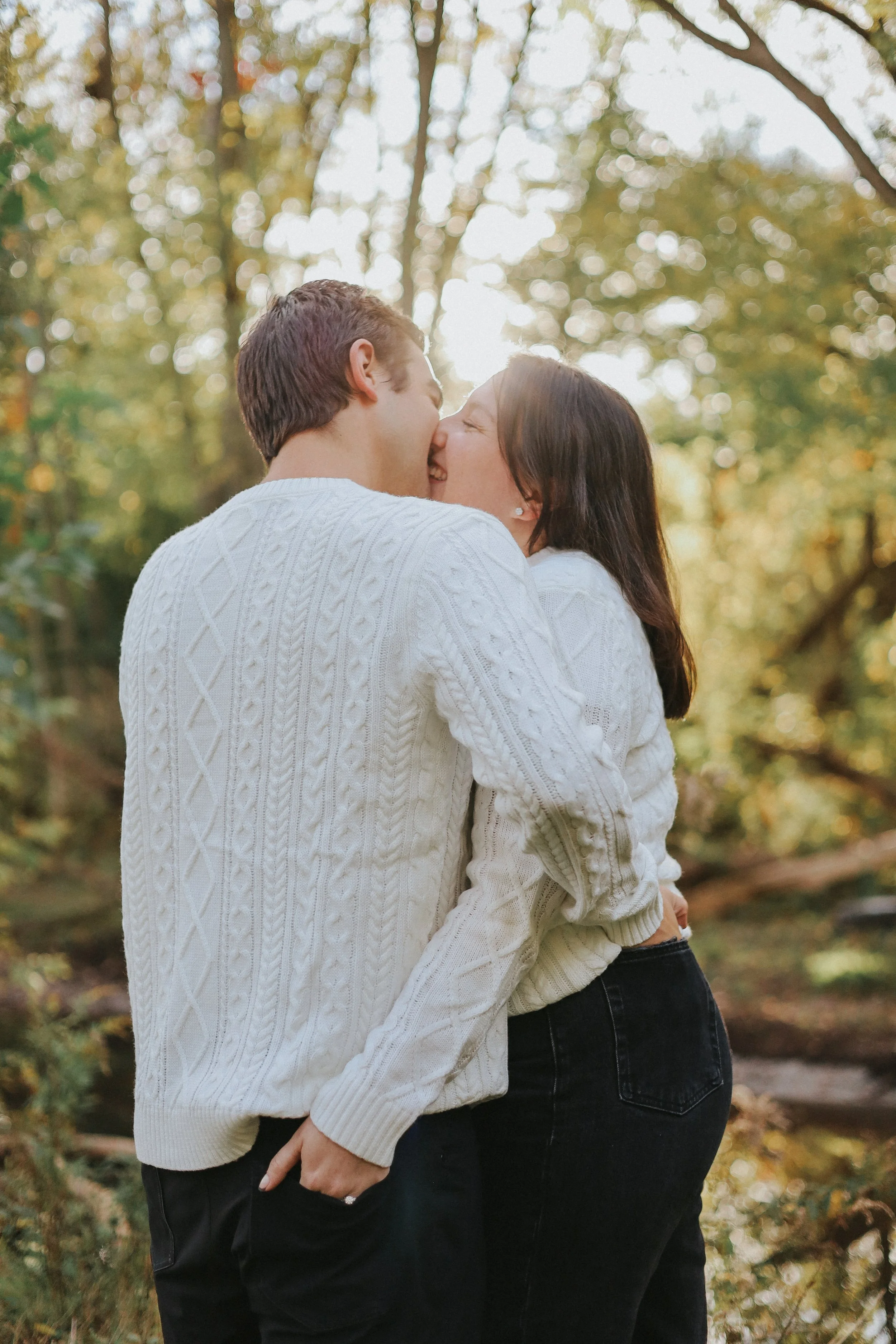 A couple hugging outdoors in a park with fall foliage, their noses touching and faces close together, both wearing white sweaters.