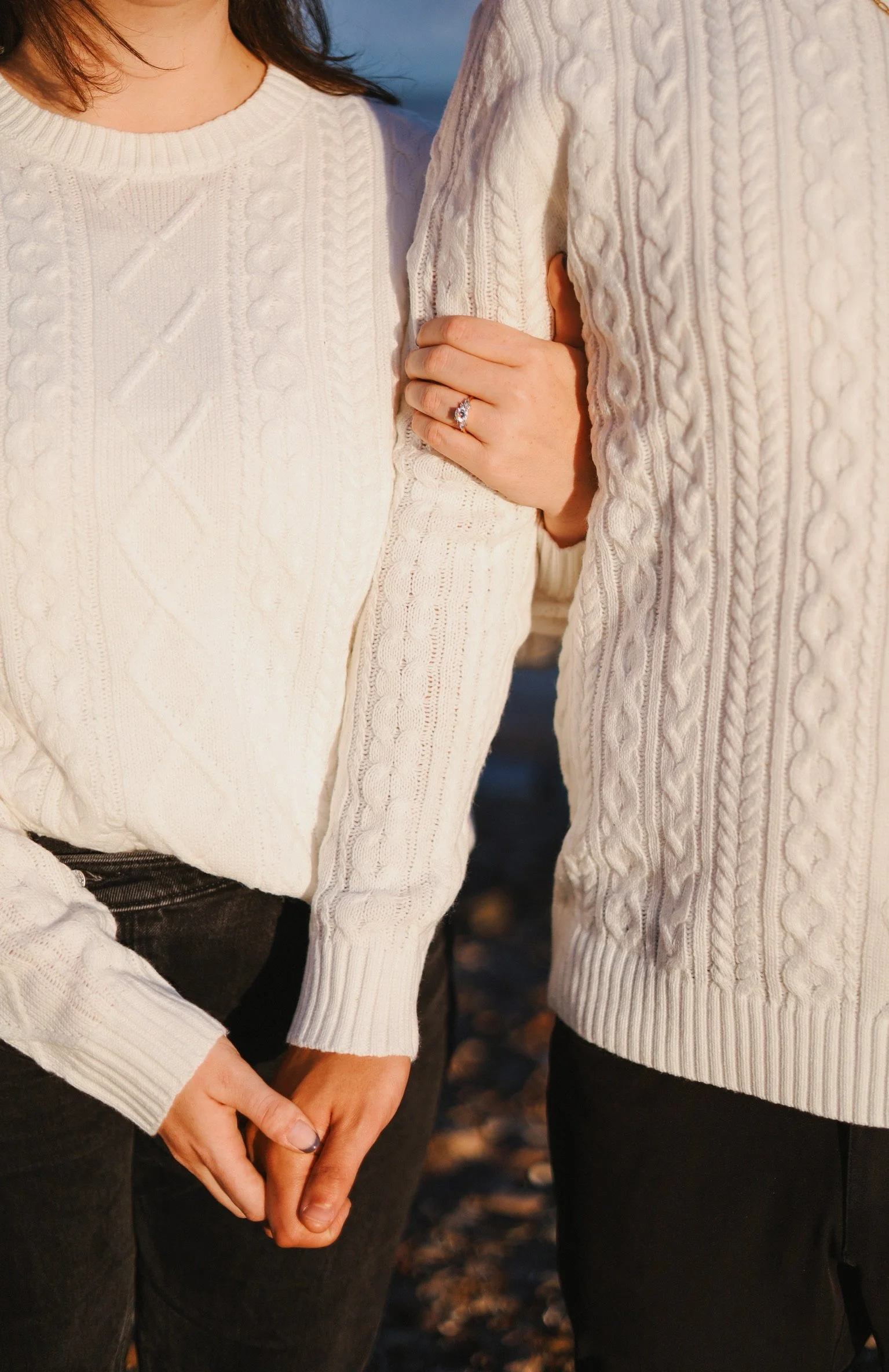 Close-up of two women wearing cream-colored cable-knit sweaters, one with a ring on her finger, holding hands.