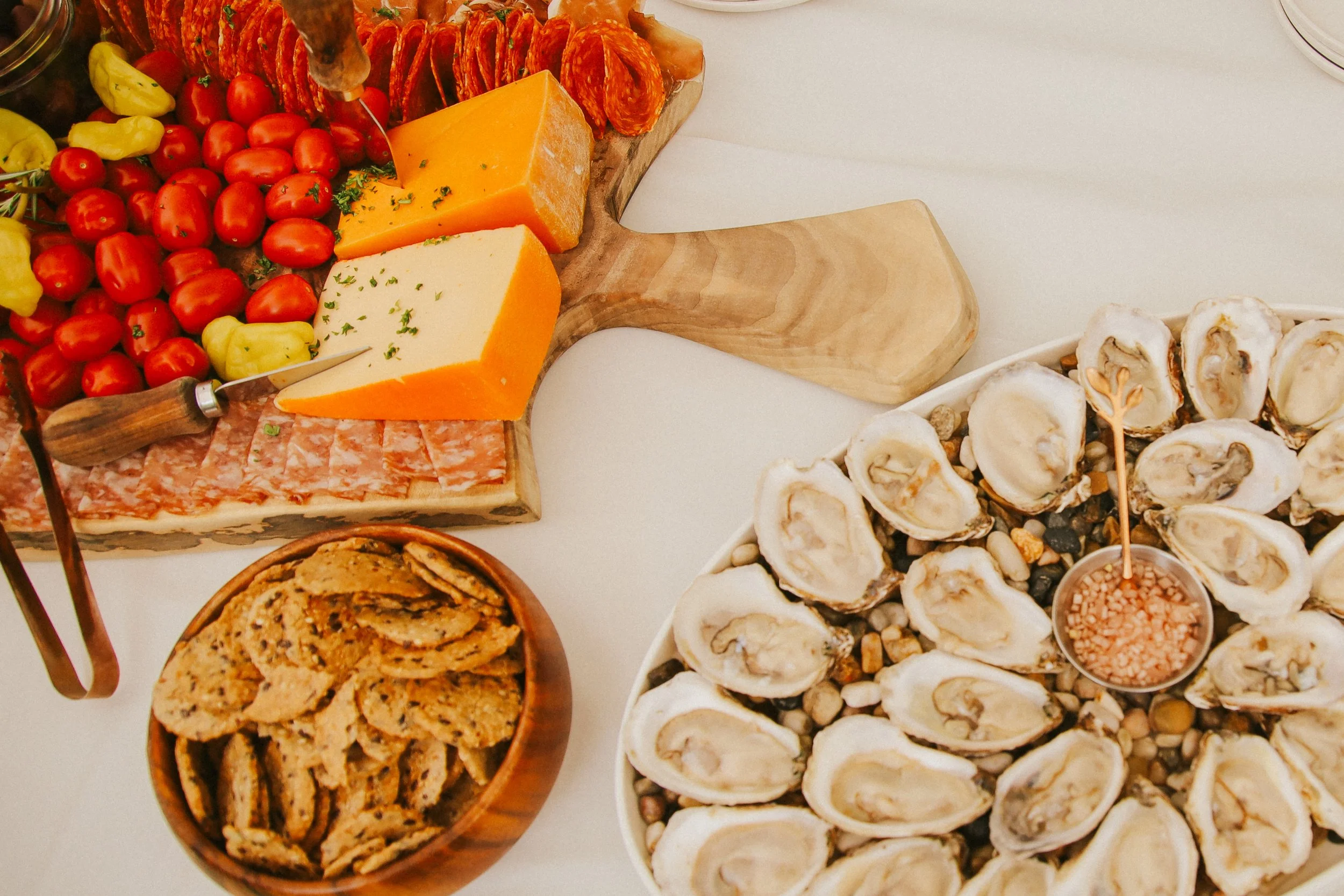Cheese and cured meats on a wooden serving board, a bowl of cherry tomatoes and bell peppers, a plate of sliced cookies, and a tray of oysters on a bed of rocks with a small dish of sauce.