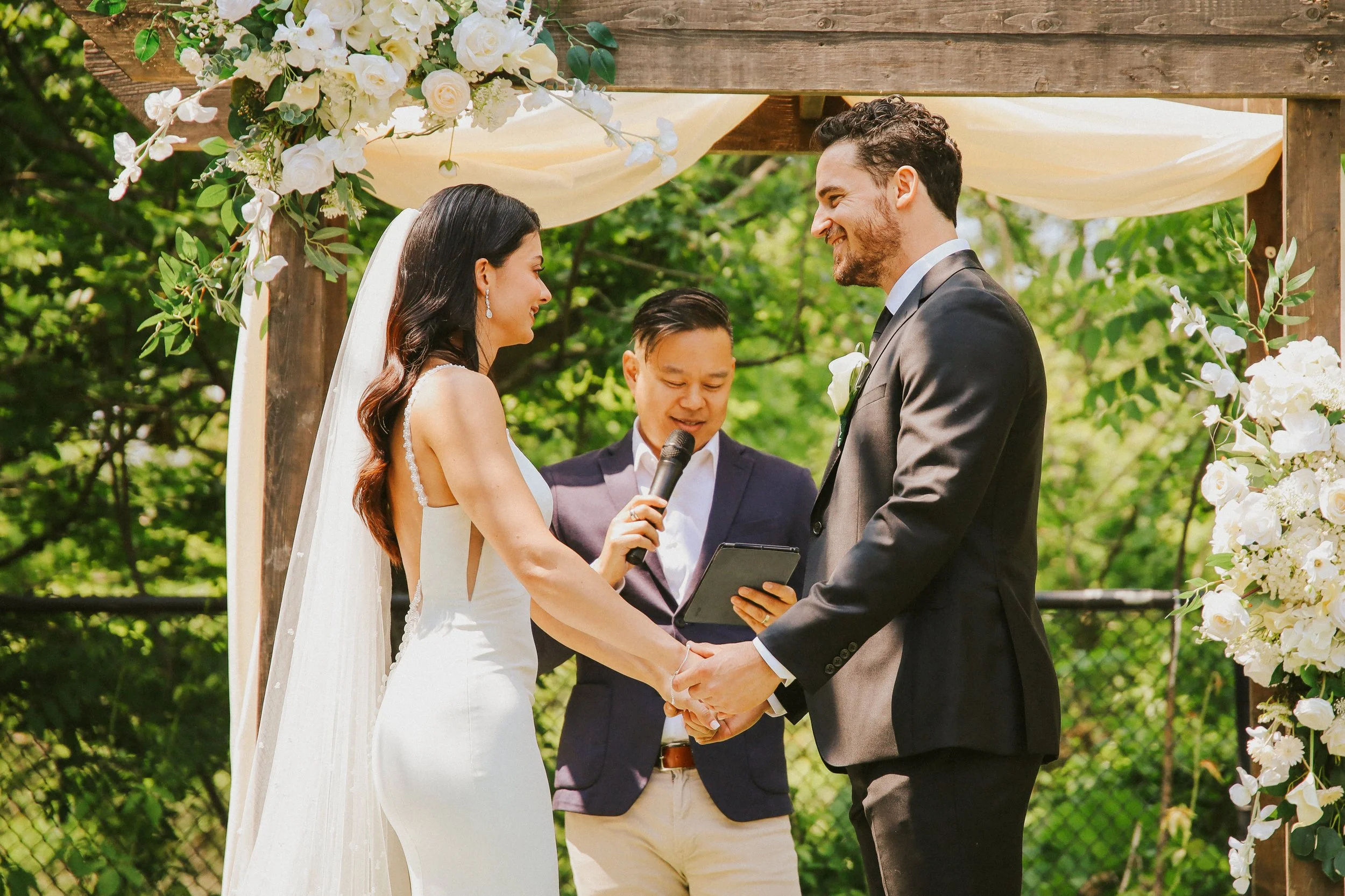 A bride and groom exchanging vows at an outdoor wedding ceremony. They are holding hands and smiling at each other, with a officiant standing between them. The ceremony is decorated with floral arrangements and greenery.