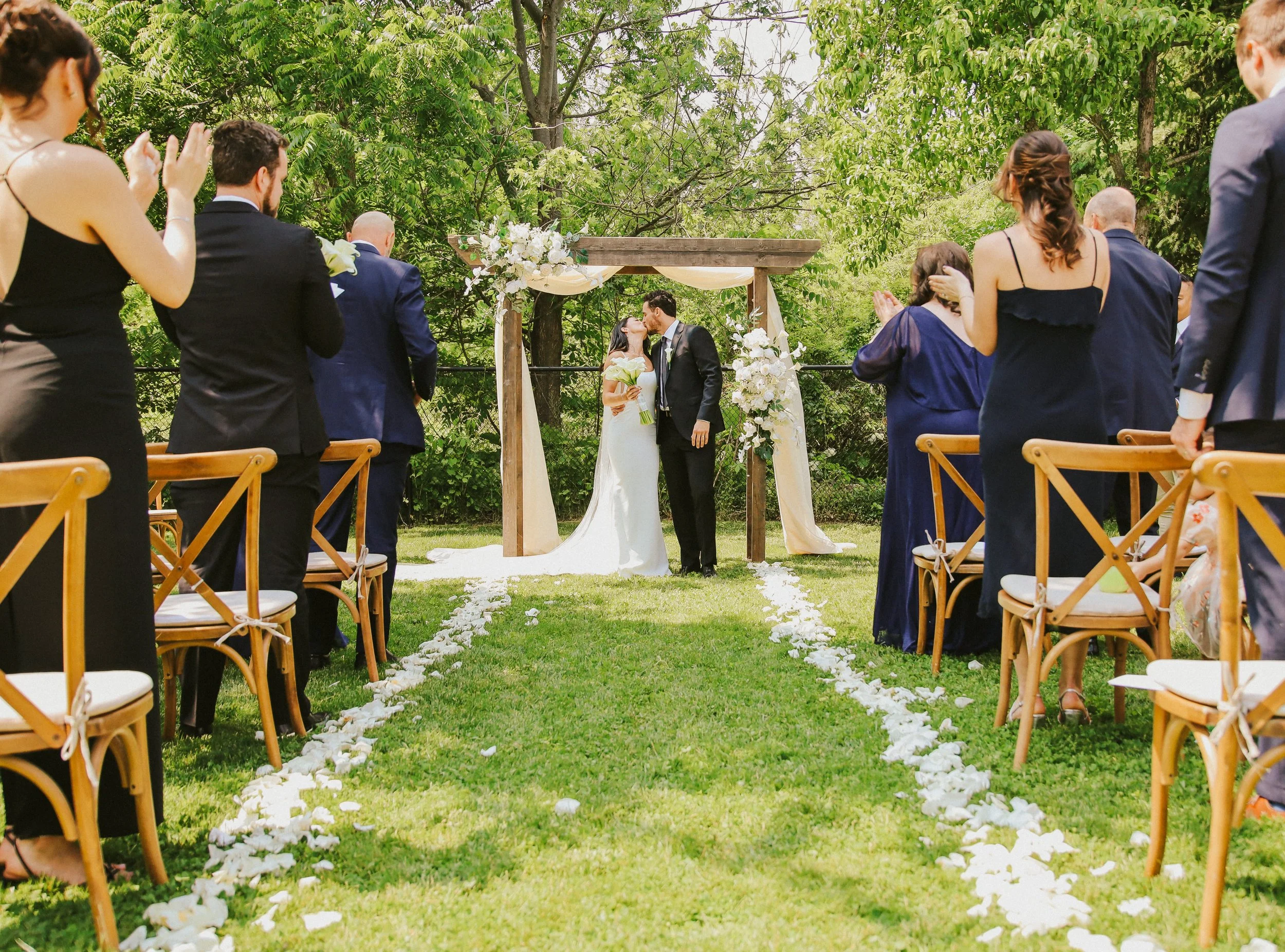 A wedding ceremony taking place outdoors in a garden, with a bride and groom kissing under a wooden arch decorated with flowers, surrounded by seated guests and lush green trees.