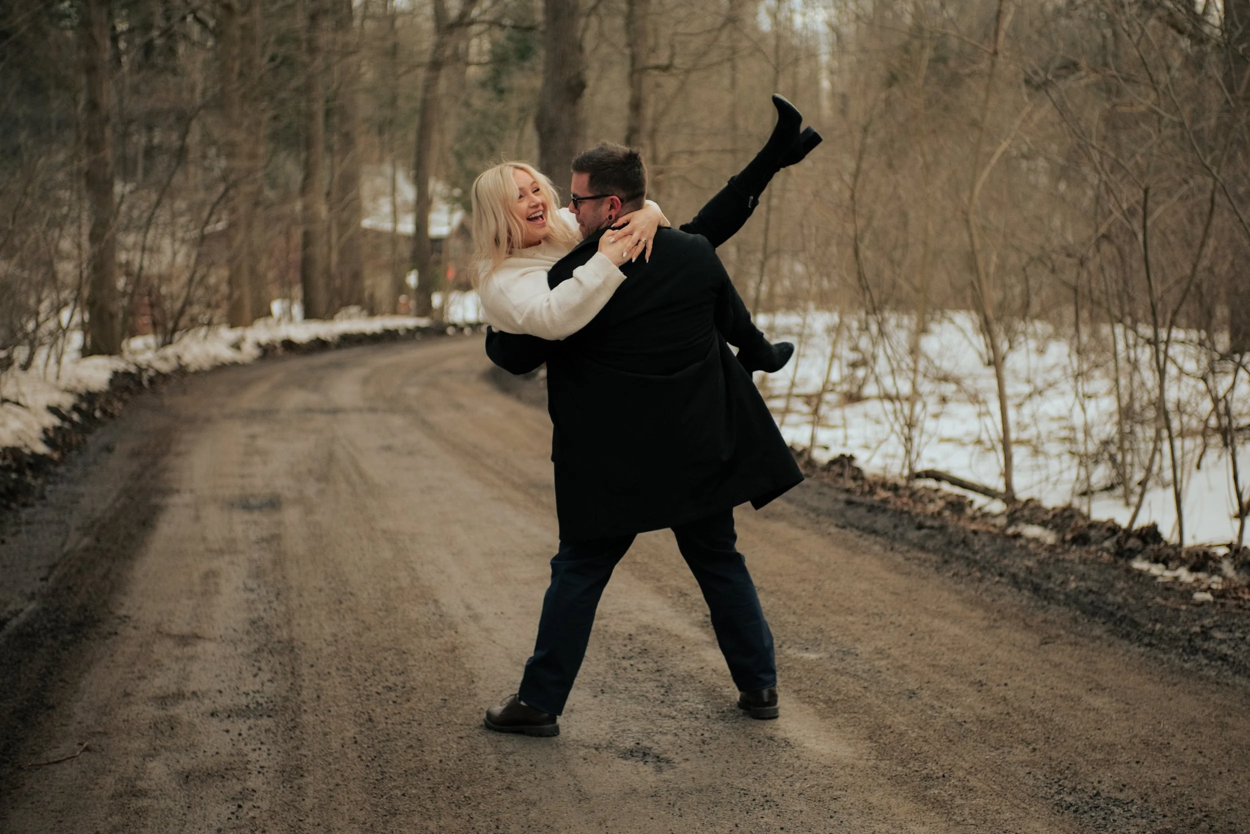 A man in dark clothing carrying a woman in a white coat on a dirt path in a wooded area, with some snow on the ground, during late fall or winter.