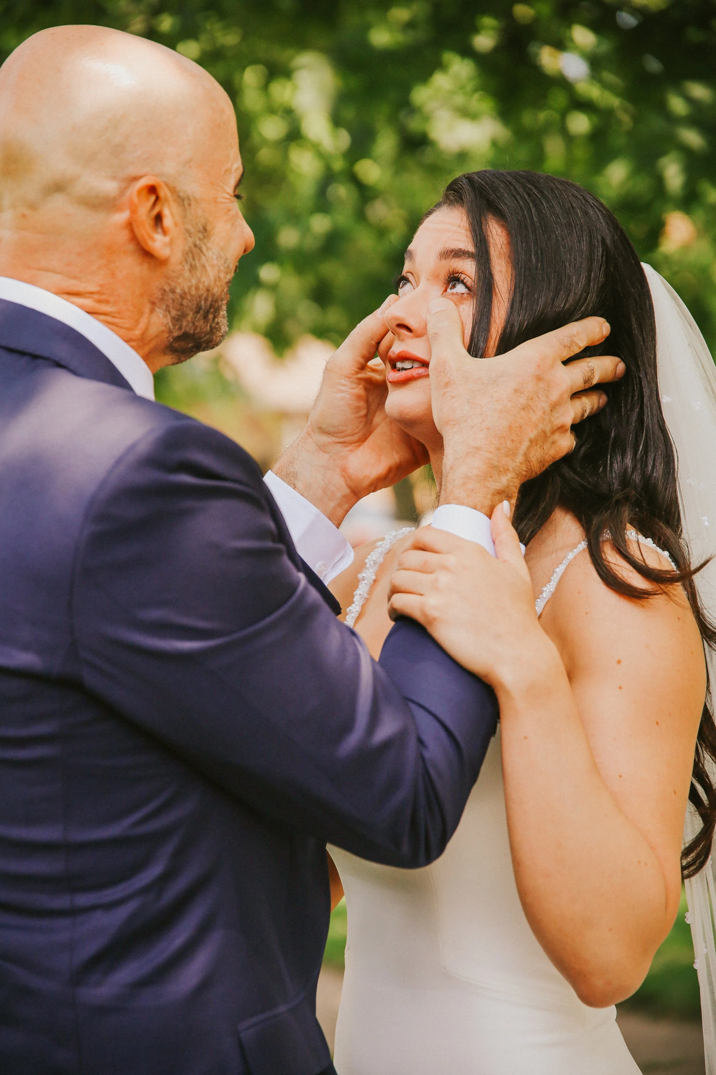 A man in a dark suit is holding a woman's face as they share an emotional moment outdoors, with green trees in the background.