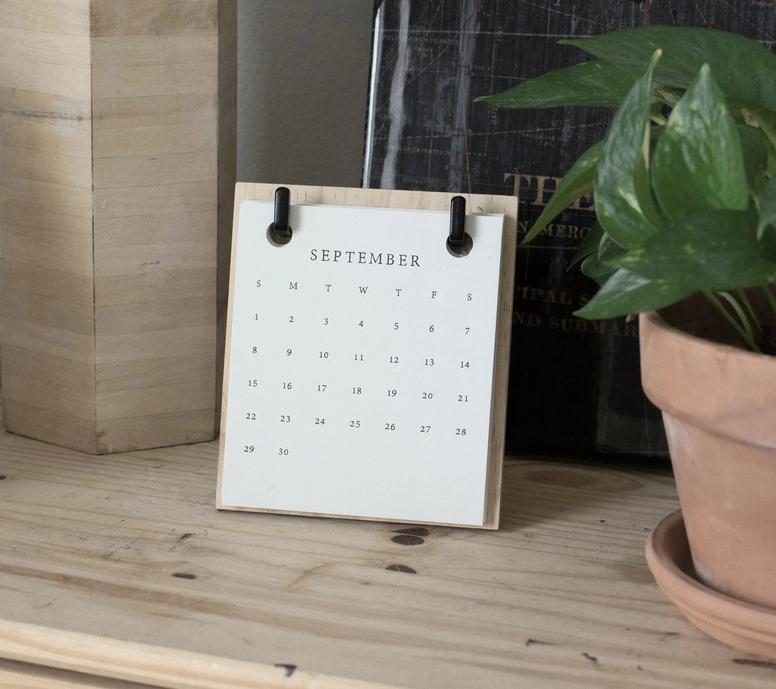 A wooden desk with a small calendar displaying the month of September, a potted plant with green leaves, and a dark surface in the background.
