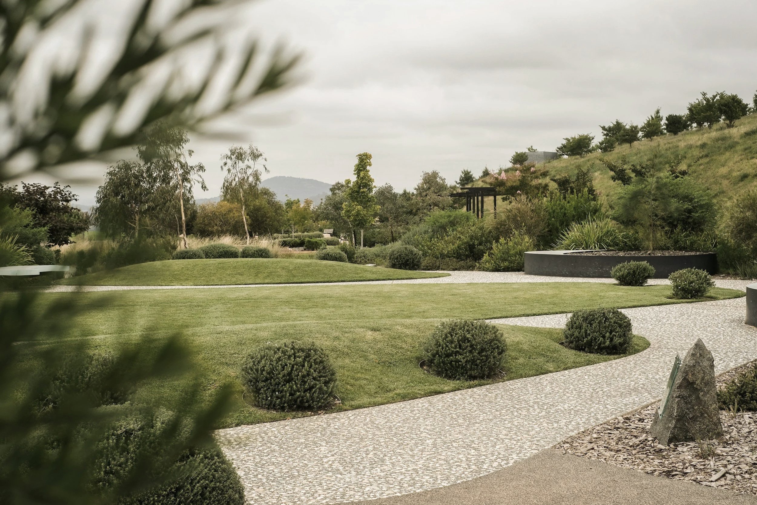 A landscaped garden with a lawn, trees, shrubs, stone pathways, and hilly terrain in the background under a cloudy sky.