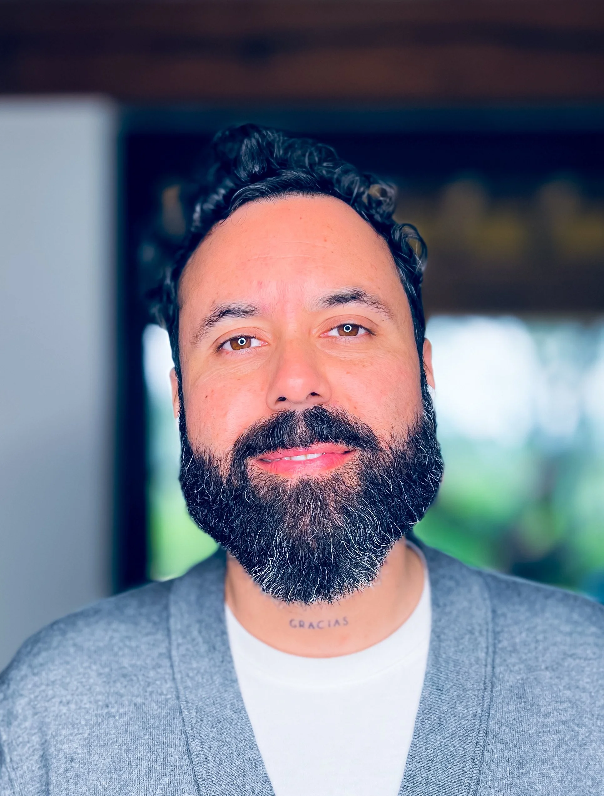 Close-up of a man with dark, curly hair and a full beard, wearing a gray cardigan over a white shirt. The background is blurred, showing an indoor setting with natural light, and the word 'GRACIAS' tattooed on his neck.