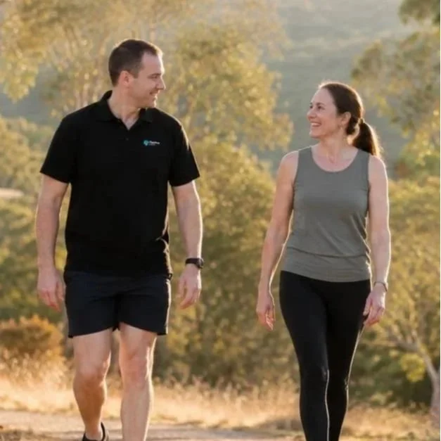 A man and woman walking together outdoors in a park during daytime, smiling at each other.