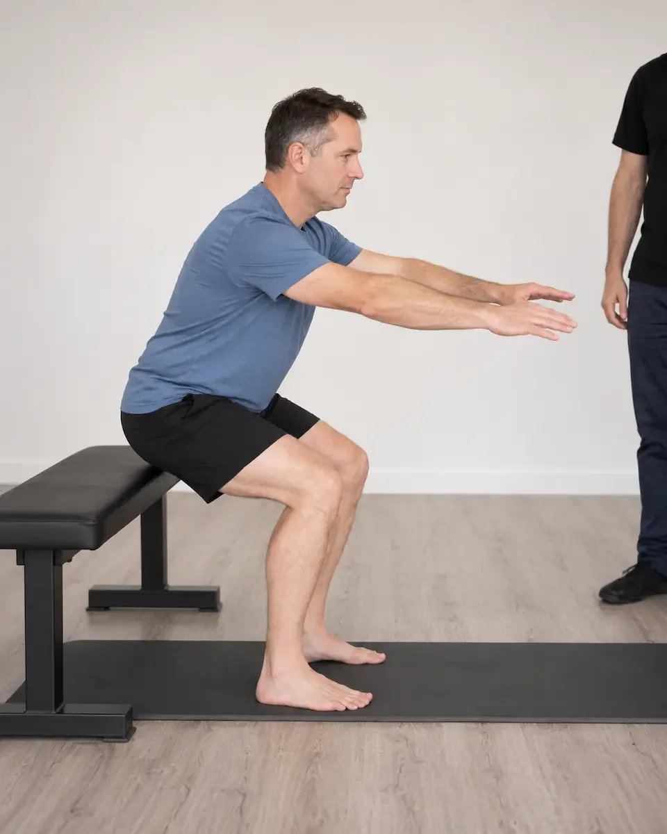 Man practising a sit-to-stand exercise from a bench with a clinician nearby.