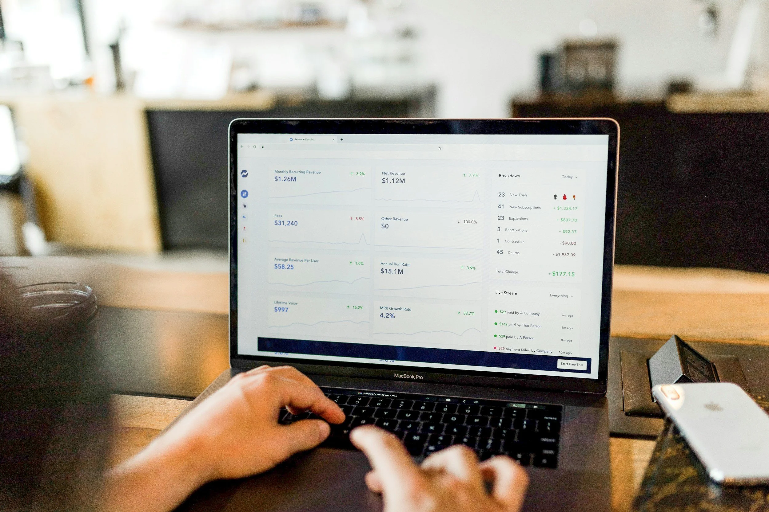 A person using a MacBook Pro laptop on a wooden table in a cafe or coworking space. The laptop screen displays a revenue dashboard with charts and financial data. In the background, there are blurred shelves and other furniture.