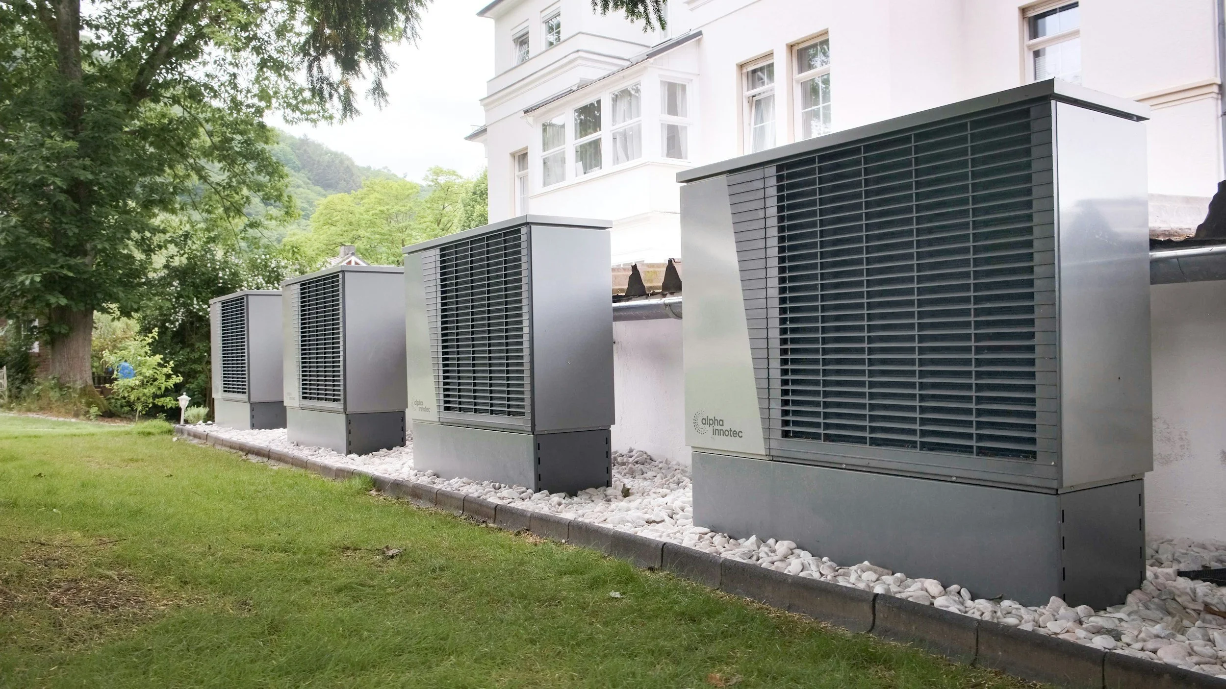 Four large outdoor air conditioning units or heat pumps installed along the side of a white residential building, with a landscaped yard and trees nearby.
