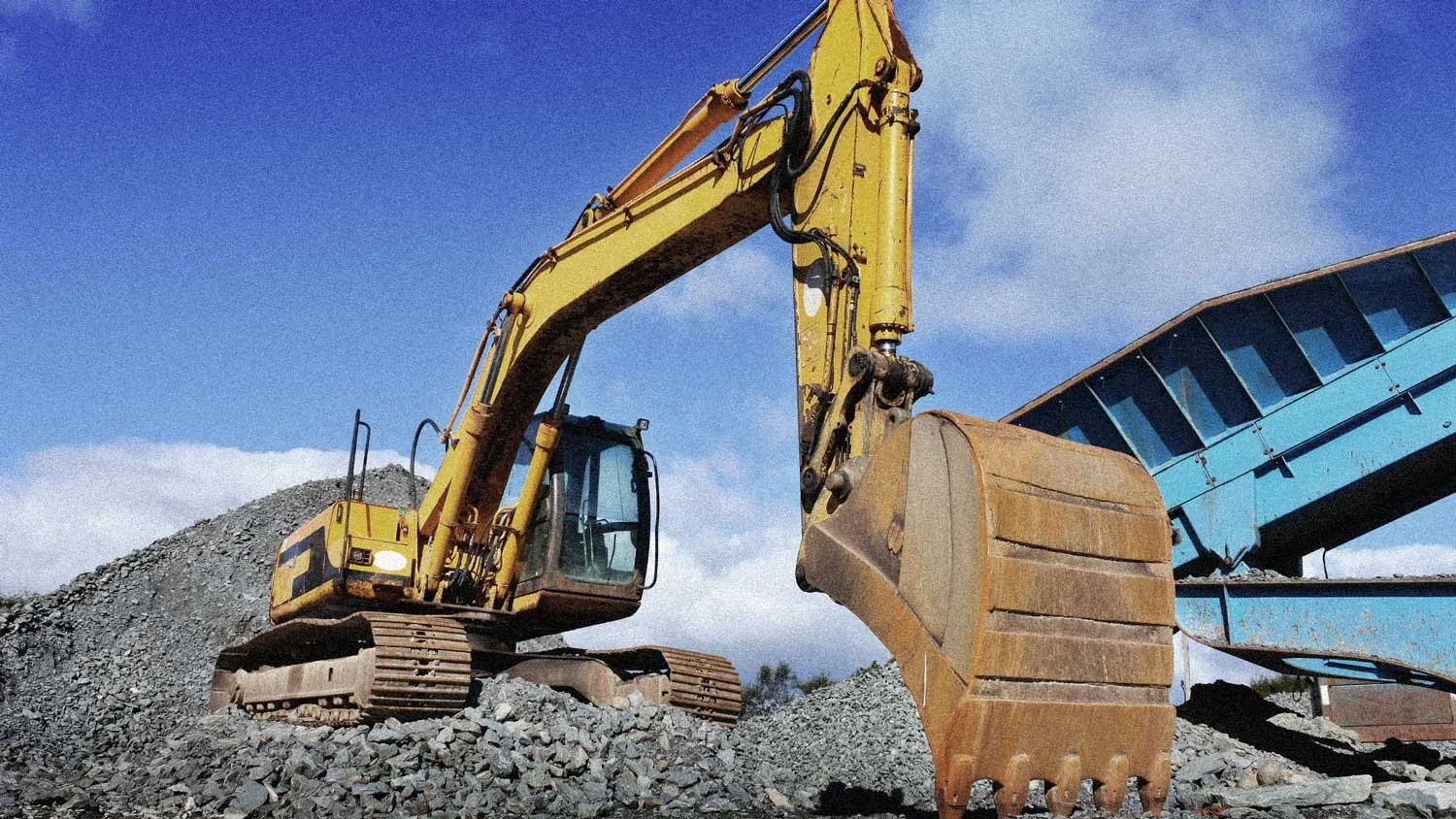 A yellow excavator working on a rocky construction site with a blue conveyor belt and a cloudy sky in the background.