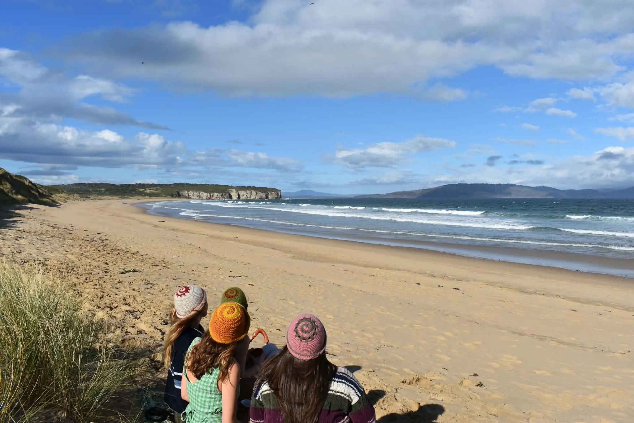 A group of people sitting on the sandy beach, facing the ocean, wearing colorful knitted hats, with cliffs and distant land visible under a partly cloudy sky.
