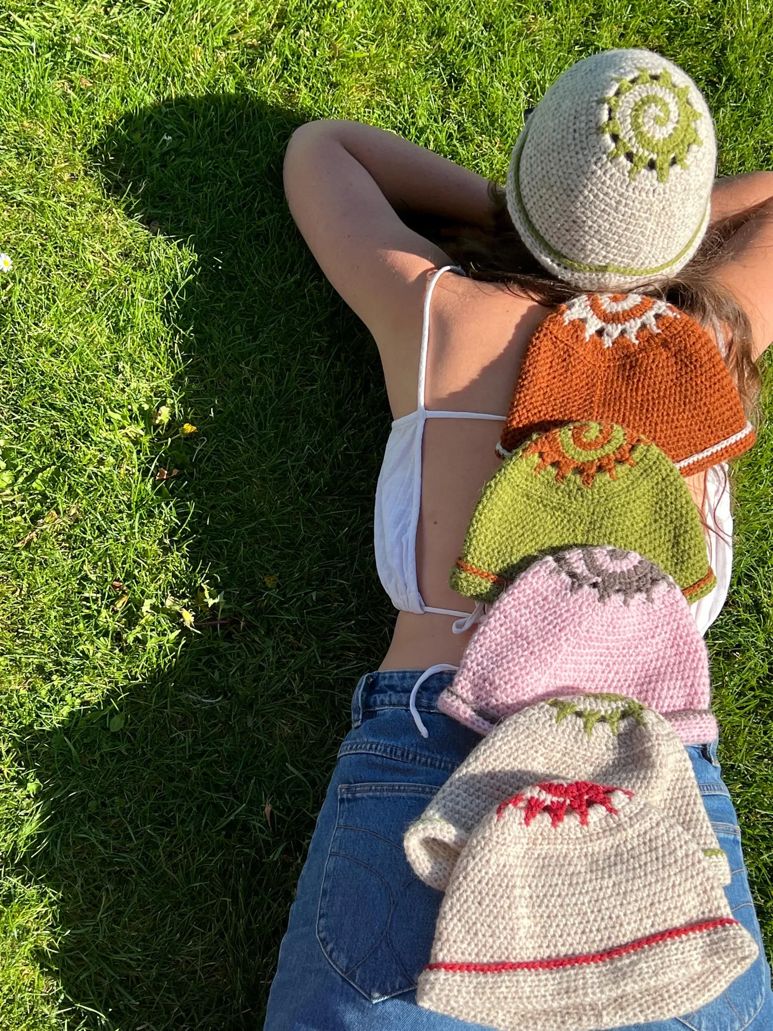 A person lying on their back on green grass, wearing a white spaghetti strap top, a knitted hat with a green sun motif, and blue jeans. Several knitted hats in different colors are placed along their body.