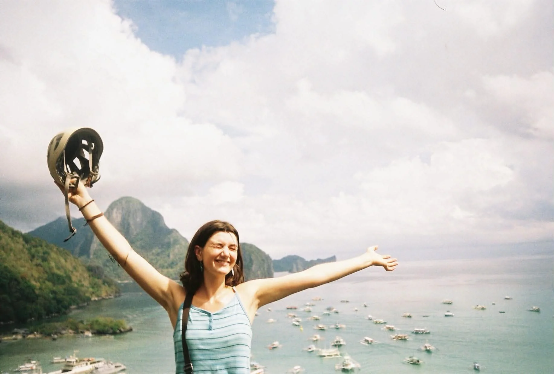 A woman with arms outstretched smiling, holding a helmet in her hand, with a coastal landscape, boats in the water, and mountains in the background.