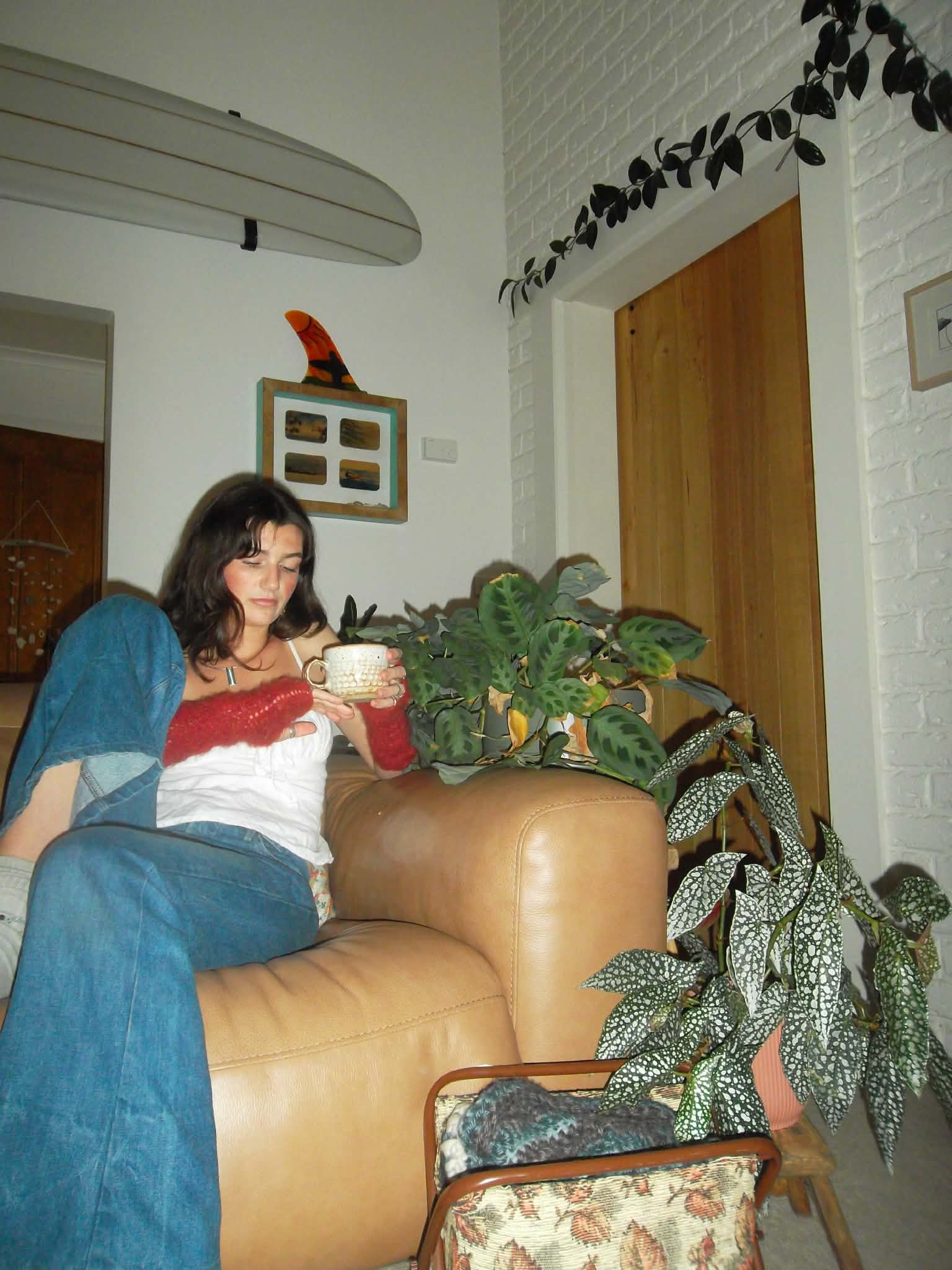 A woman with dark wavy hair sitting on a couch, holding a mug, in a room with potted plants, a white brick wall, a wooden door, and some framed pictures.