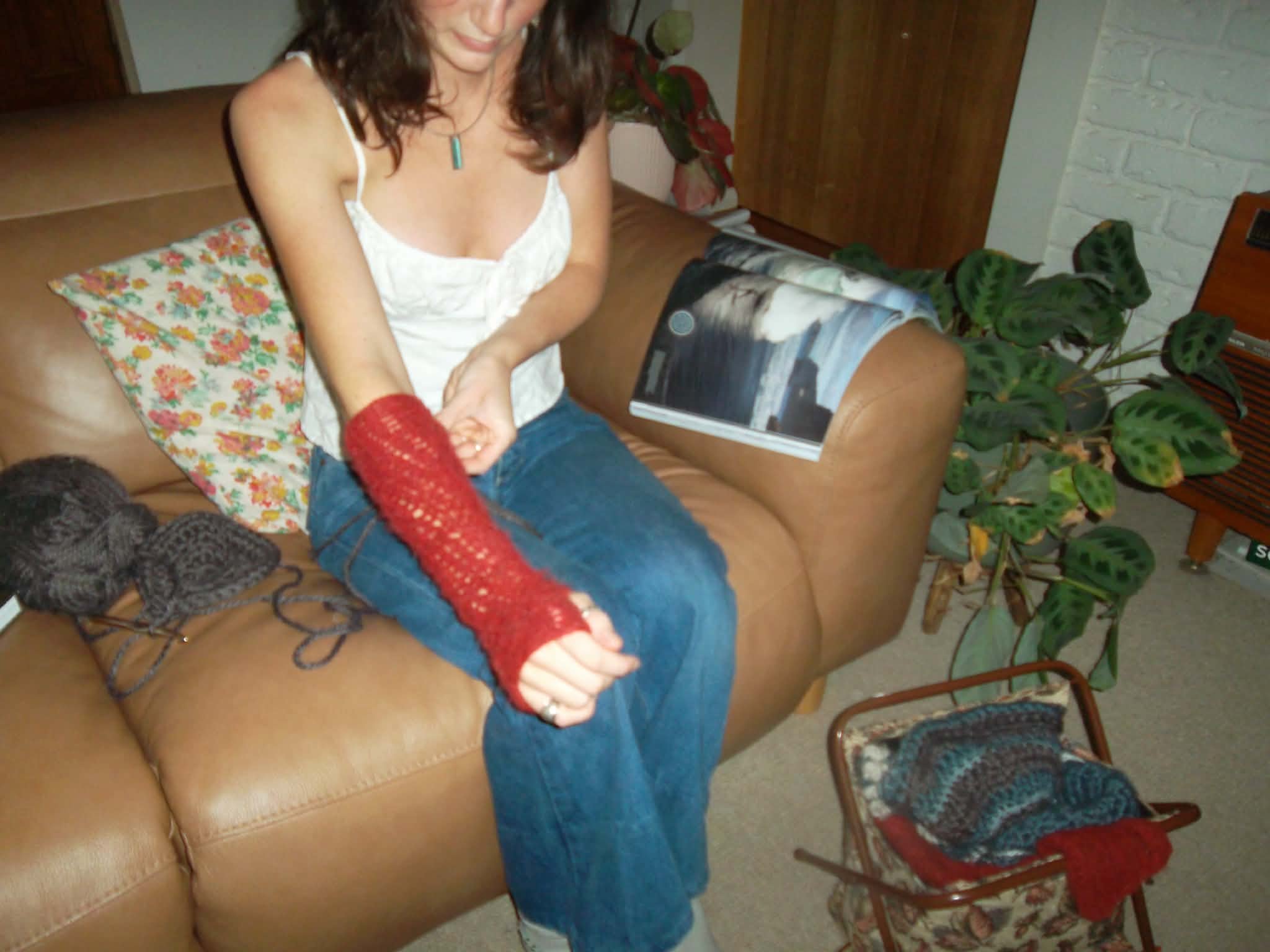 A woman with long brown hair wearing a white sleeveless top, blue jeans, and a turquoise pendant necklace sitting on a brown leather couch. She is holding a red knitting project in her hands. There is a floral pillow behind her and a magazine with a photograph of a house on her lap. To her right, there is a large leafy houseplant, a wooden chair with a knitted blanket and a red bag on the floor nearby. The wall behind her is made of white brick and there is a wooden cabinet to her left.