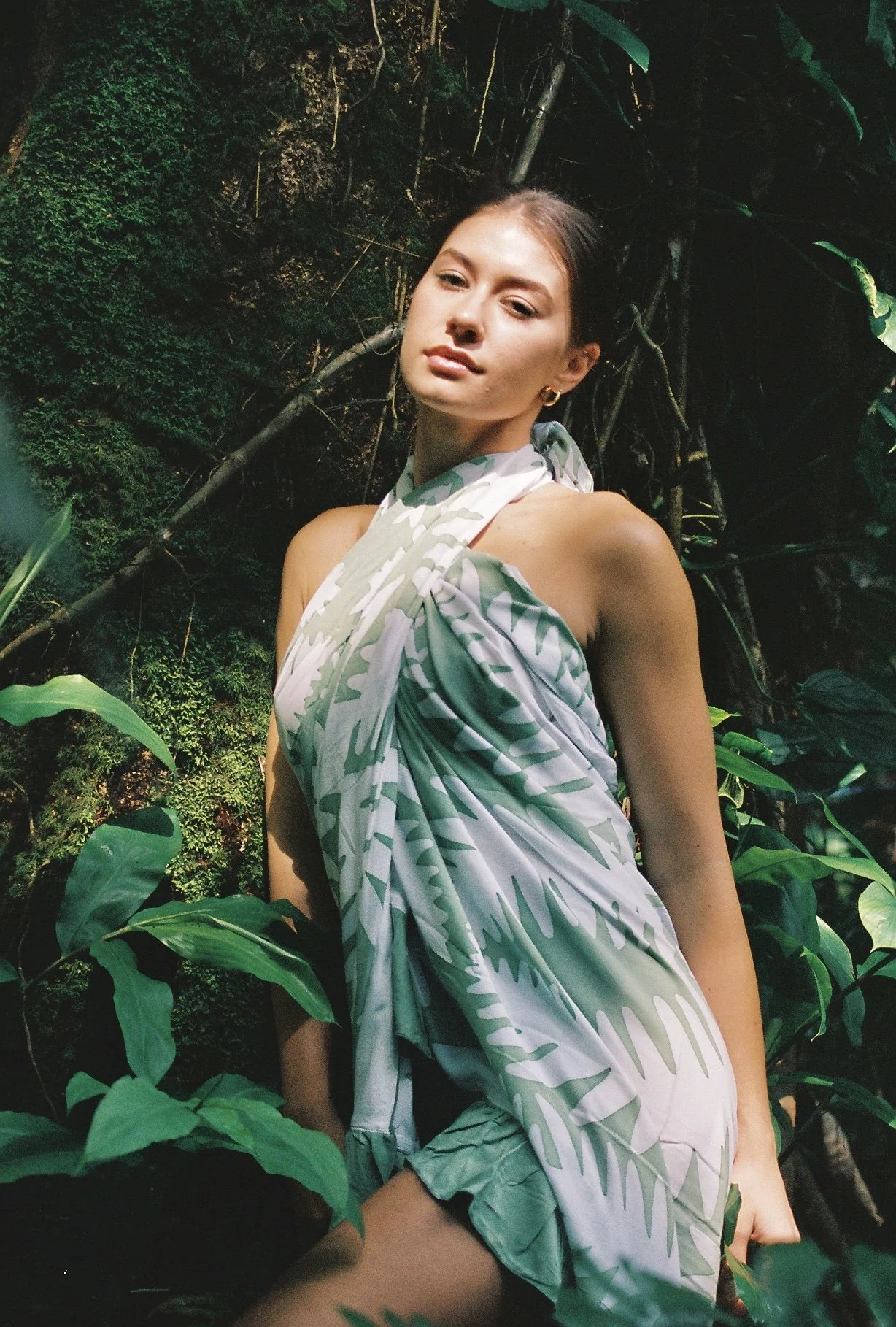 A woman with brown hair and hoop earrings posing among green plants and jungle foliage, wearing a light-colored dress with a tropical leaf pattern.