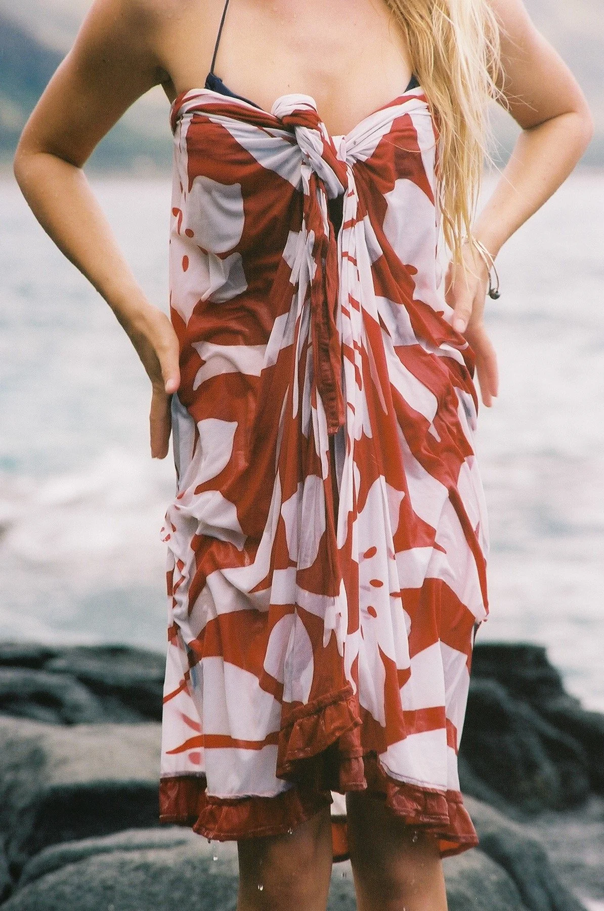 Person wearing a dress with a red and white abstract pattern, standing near water with rocks in the background.