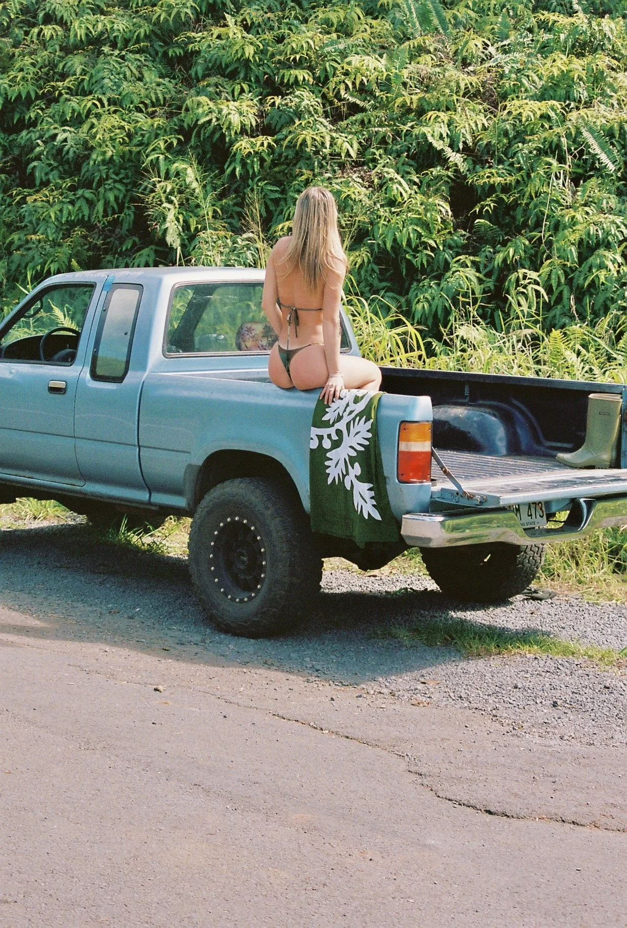 A woman in a bikini sitting on the back of a blue pickup truck parked on a dirt road with green tropical foliage in the background.
