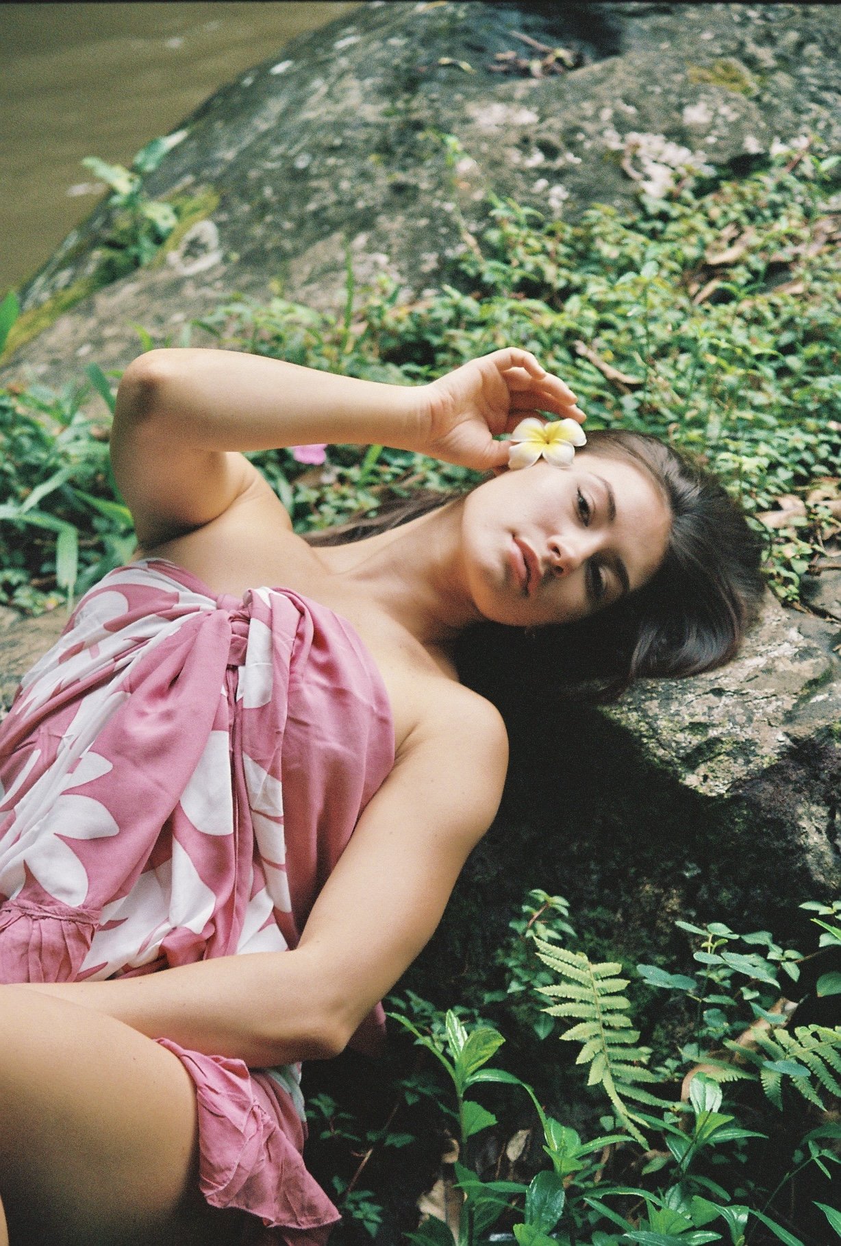 Woman lying on her side on a rock by a water body, wearing a pink and white floral dress, holding a yellow flower near her temple, surrounded by lush greenery.