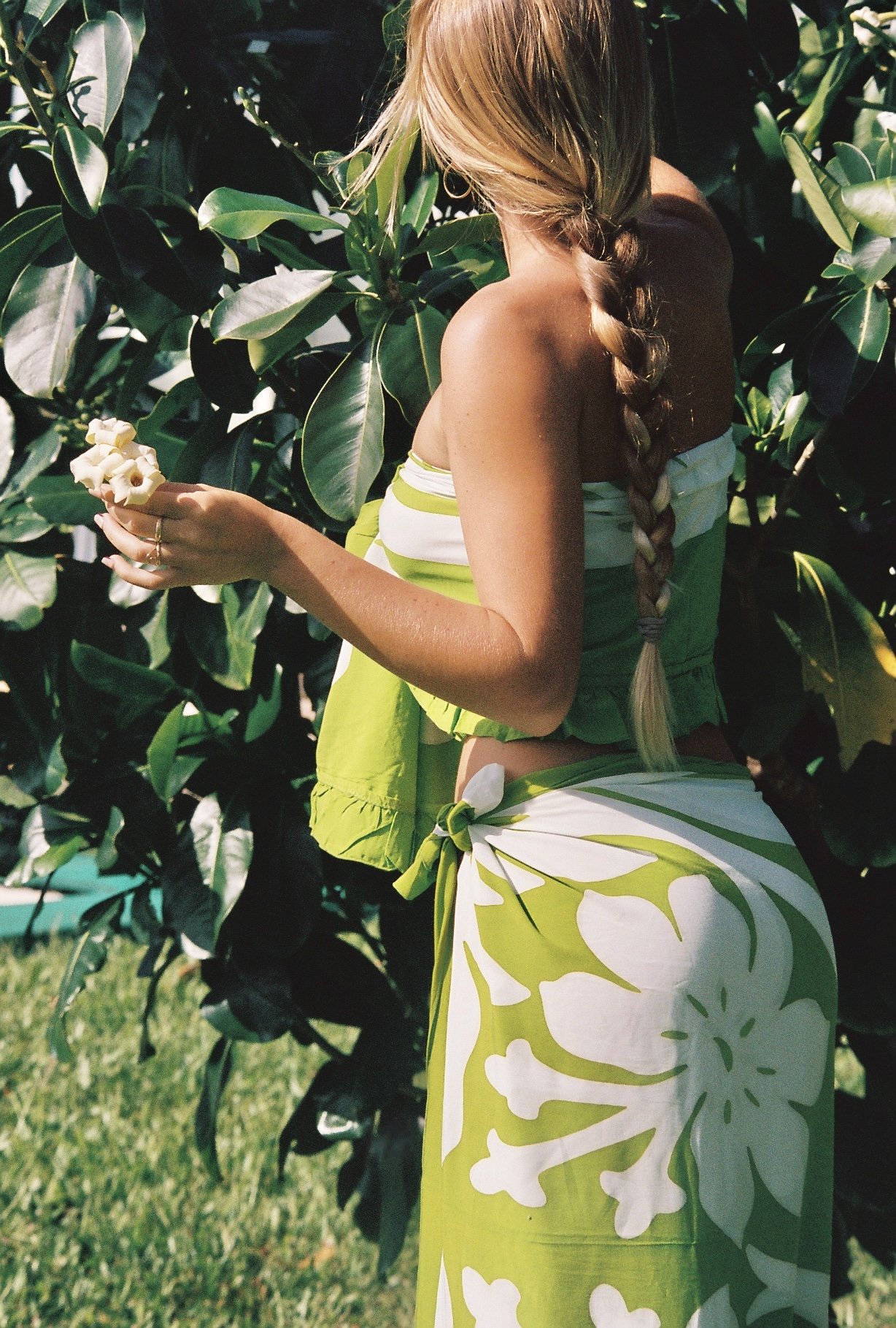 A woman with long blonde hair styled in a braid is standing outdoors among green plants. She is wearing a strapless top and a matching skirt with a green and white leafy pattern. She is holding a small bouquet of white flowers in her right hand.