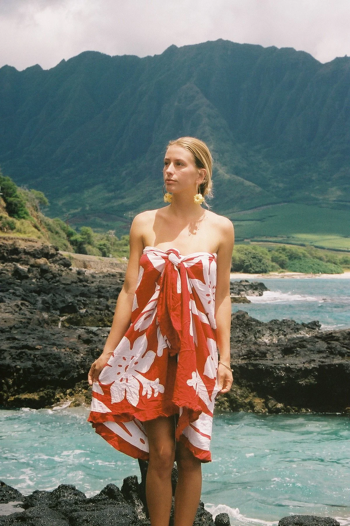 A woman standing on rocks by the ocean, wearing a red and white floral dress with a knot at the front, large yellow earrings, and a natural background of a mountainous landscape and green shoreline.