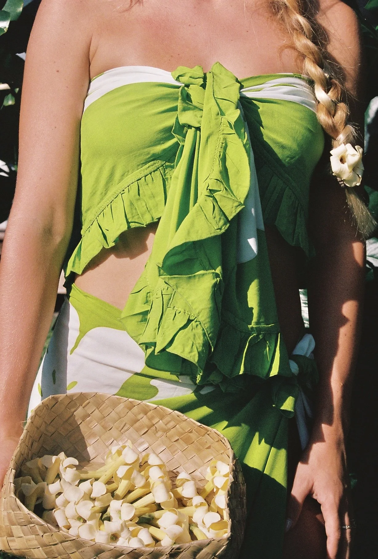 Person wearing a green and white outfit with ruffles, holding a woven basket filled with white flower buds.