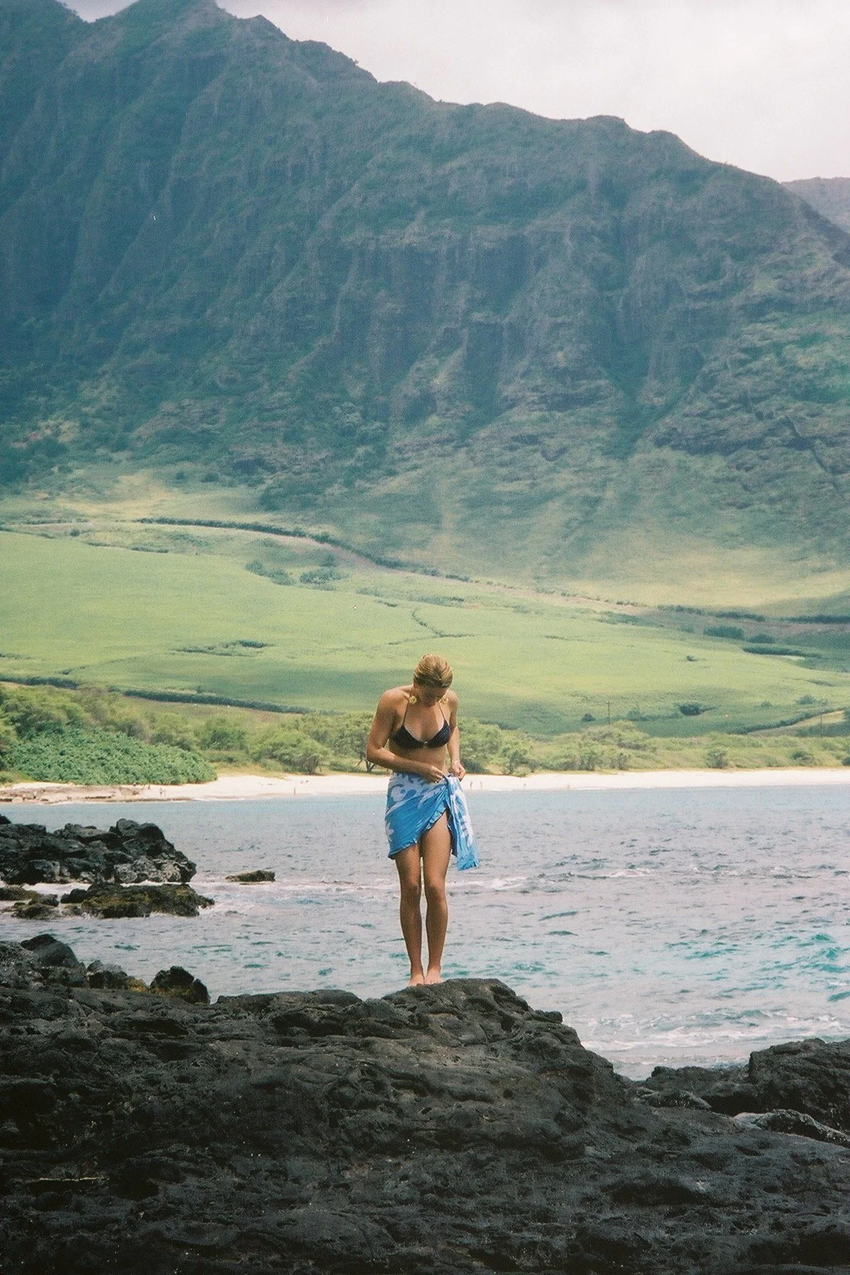 A woman standing on rocks at the beach, wearing a black bikini top and a blue sarong, with green mountains in the background and water in the foreground.