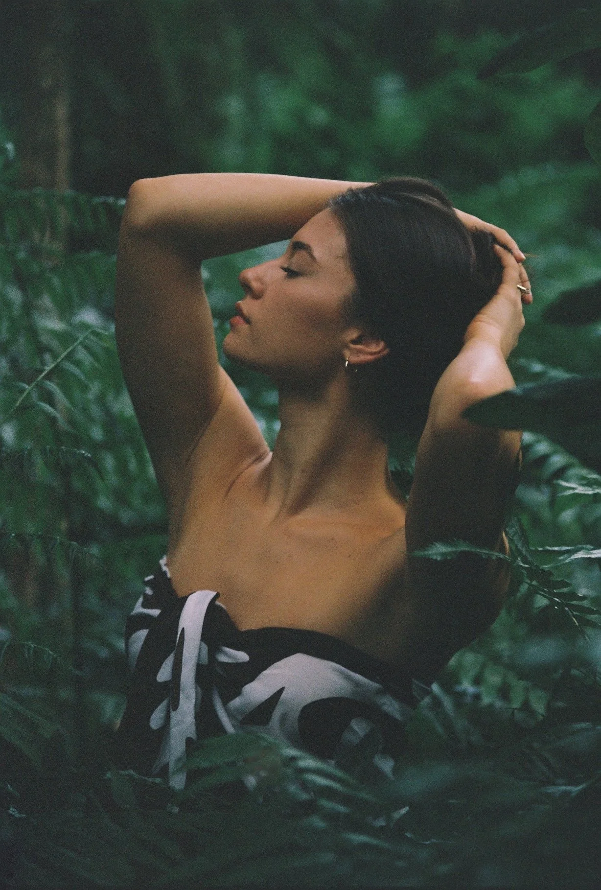 A woman with dark hair is posing with her eyes closed and hands behind her head, surrounded by lush green foliage in a jungle setting.