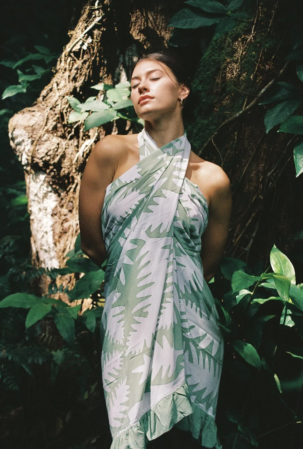 A woman with closed eyes and relaxed expression standing among dense green foliage and vines, wearing a dress with a light green and white leafy pattern.