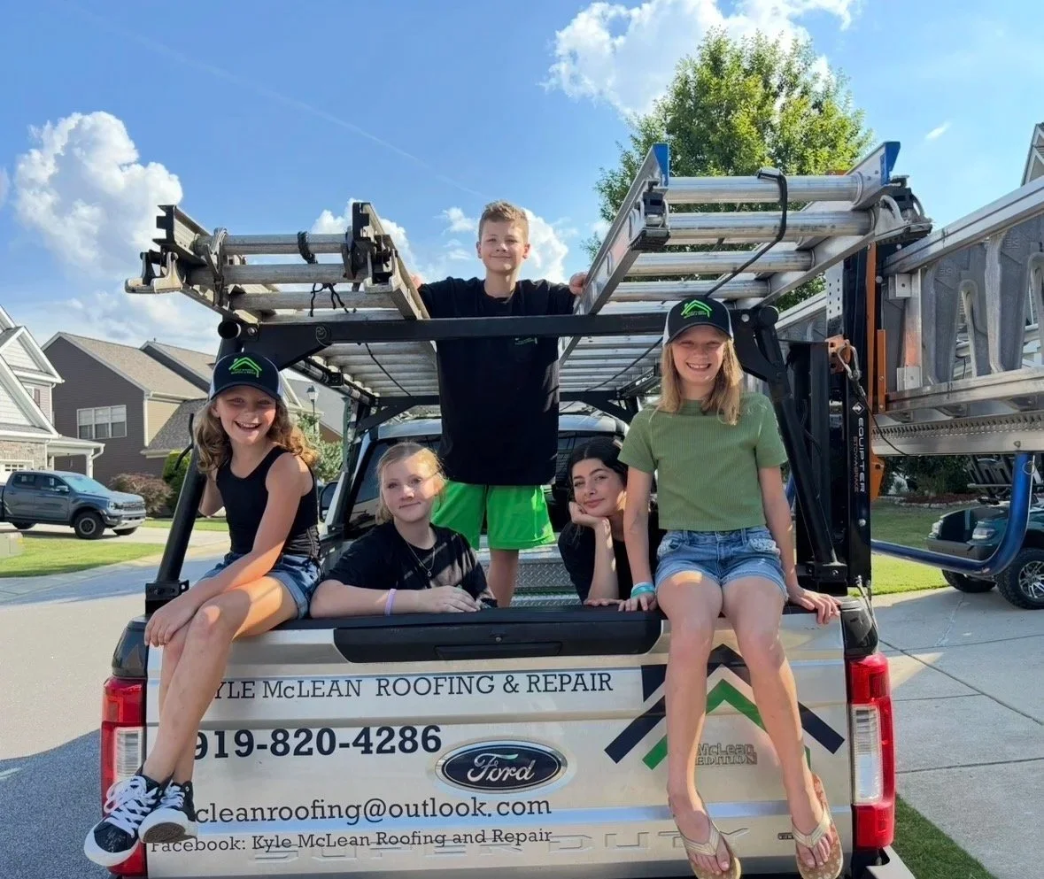 Five children sitting and kneeling on the back of a pickup truck with roofing ladders, parked in a neighborhood with houses, trees, and a blue sky.
