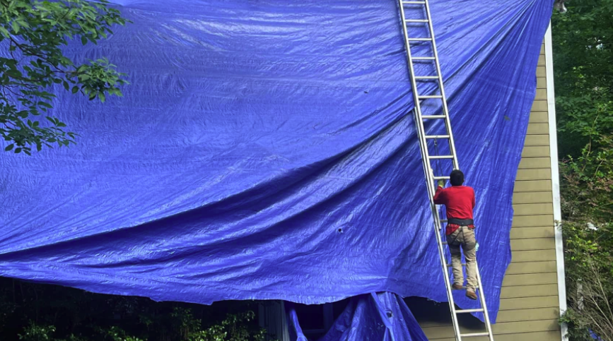 Blue tarp covering roof leak and man on ladder in red shirt