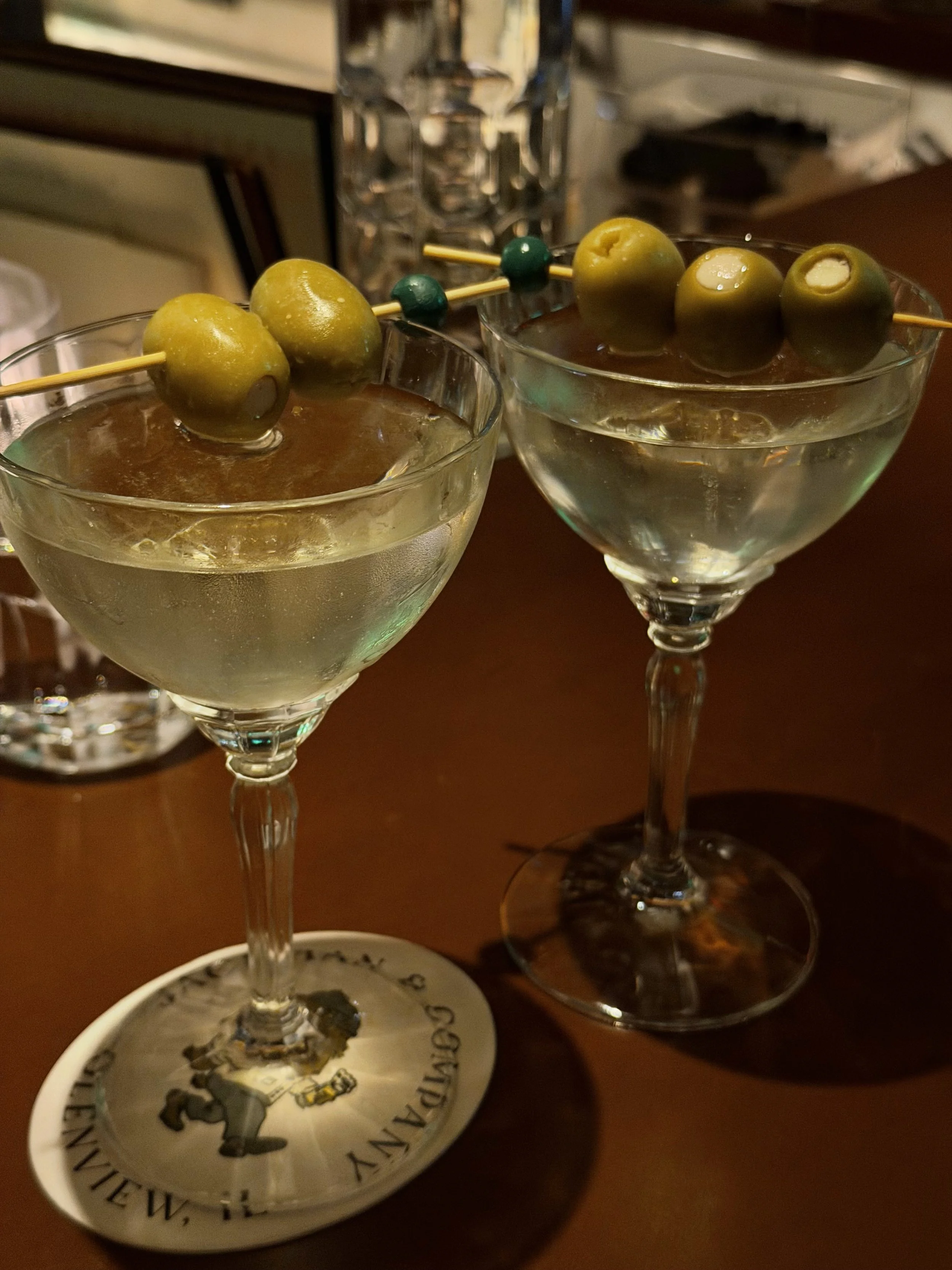 Two martini glasses filled with clear liquid and garnished with green olives on a toothpick. A decorative plate with a character and text is partially visible in front of the glasses. A glass of water and some books are in the background.
