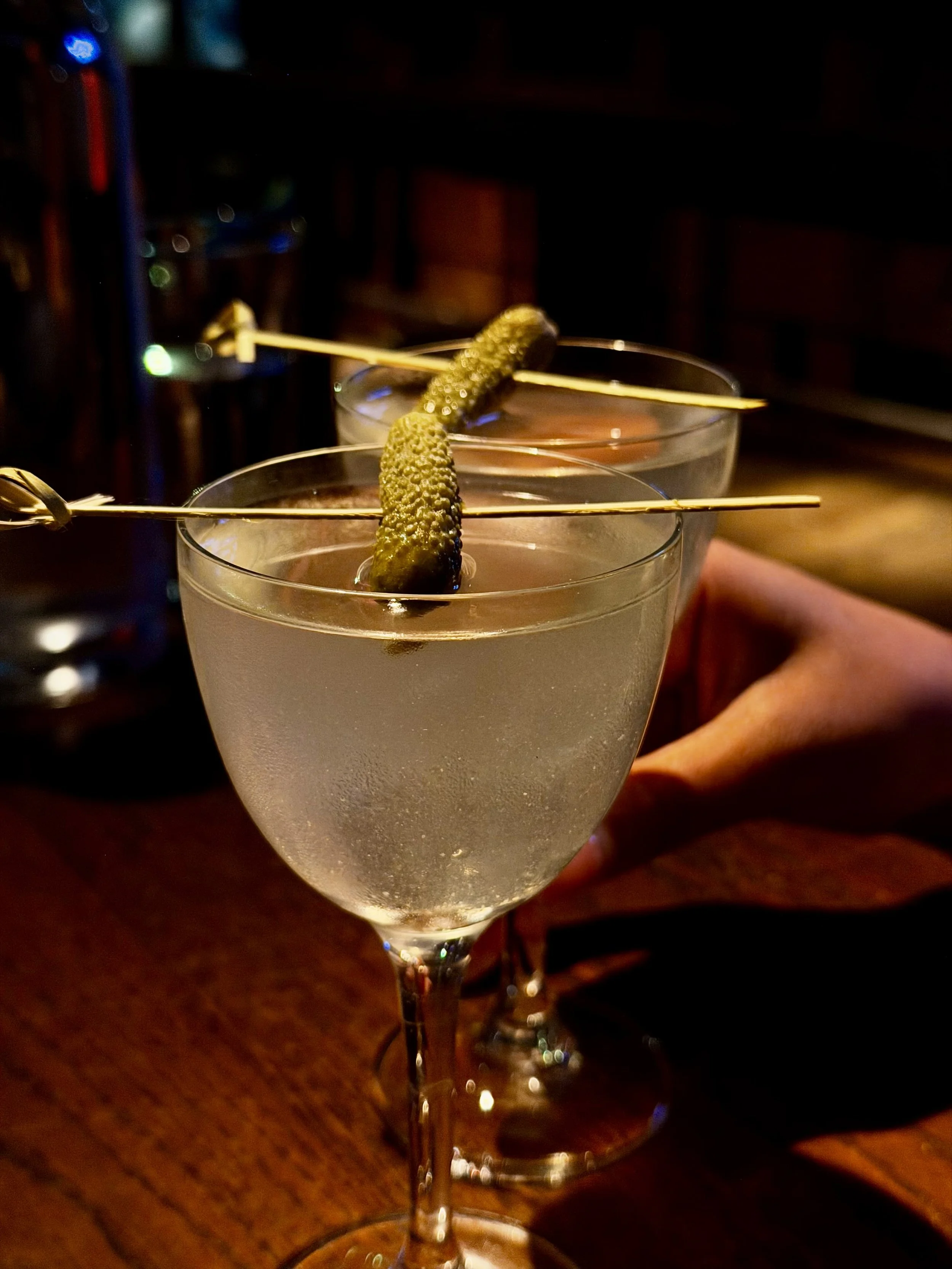 Two martini glasses with pickles on skewers, filled with a clear beverage, set on a dark wooden table in a dimly lit setting.